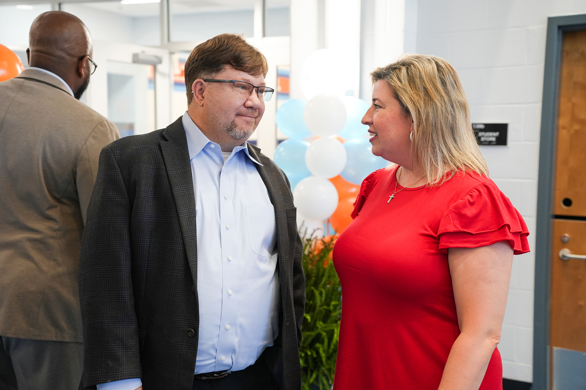 A man in a dark blazer and a woman in a red dress face each other, engaged in conversation.