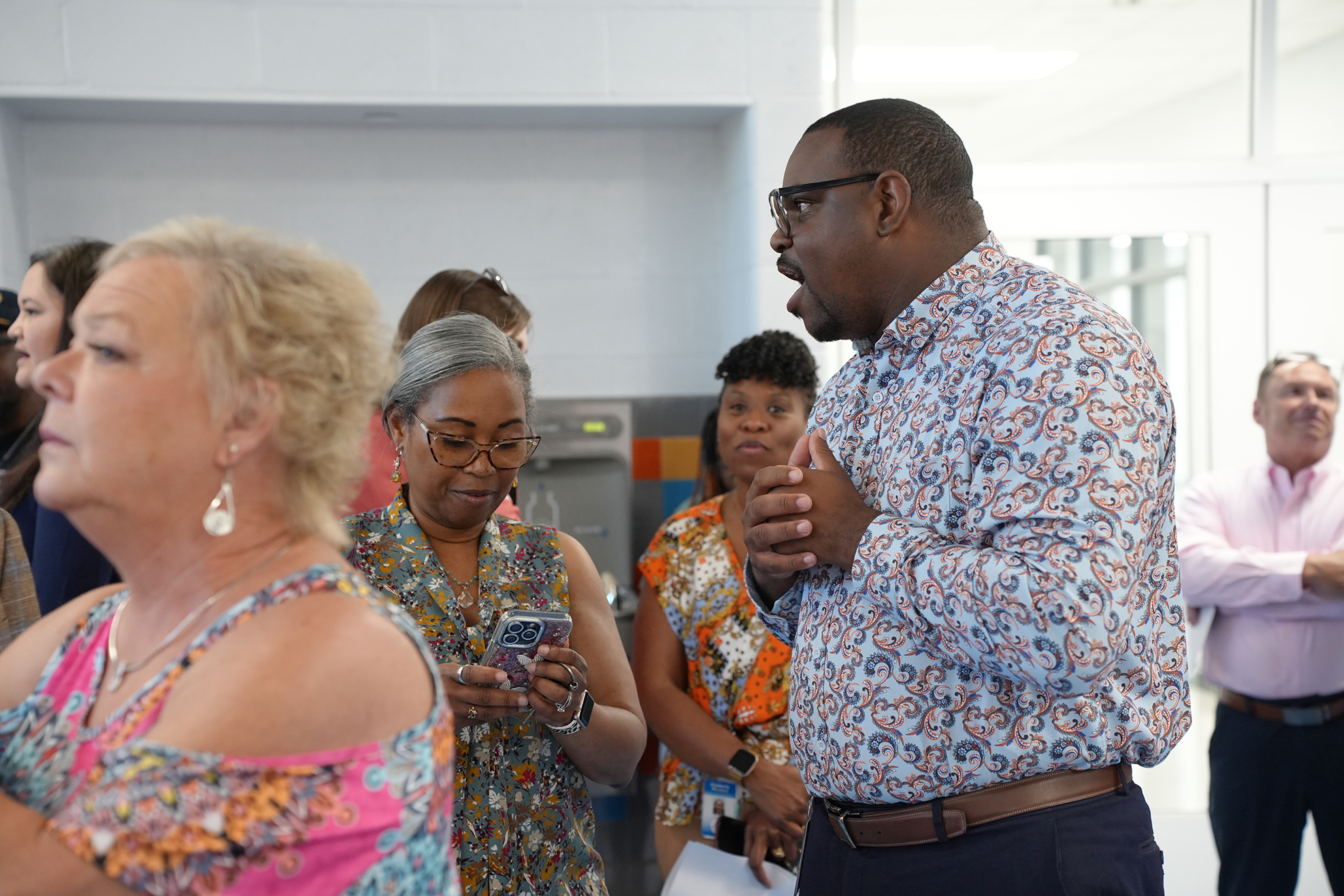 A group of people converses in a room with white walls and a white ceiling. A man wears glasses and a floral shirt.