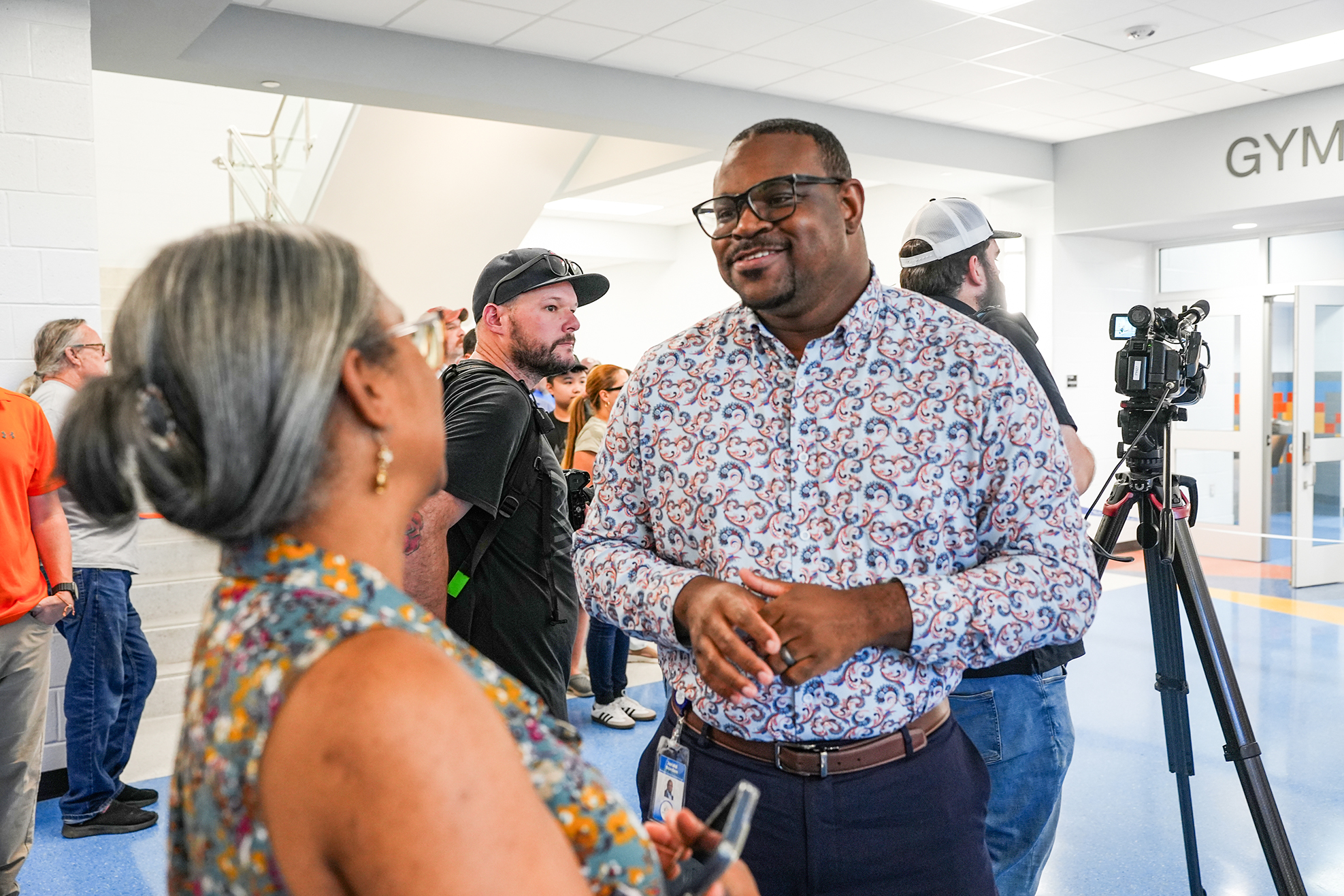 A man in glasses and a floral shirt converses with a woman in a hallway with others.