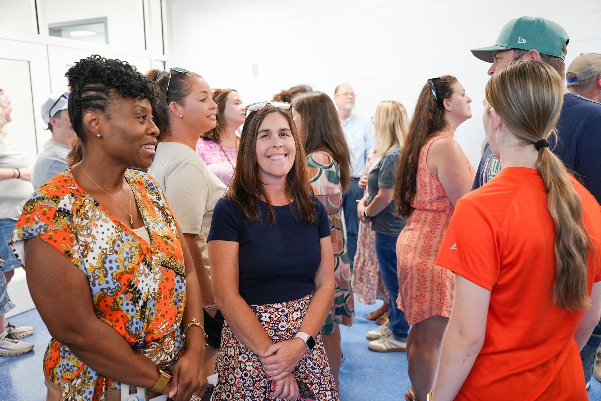 People gather indoors, wearing summer clothing, with two women in floral and blue shirts at the front.