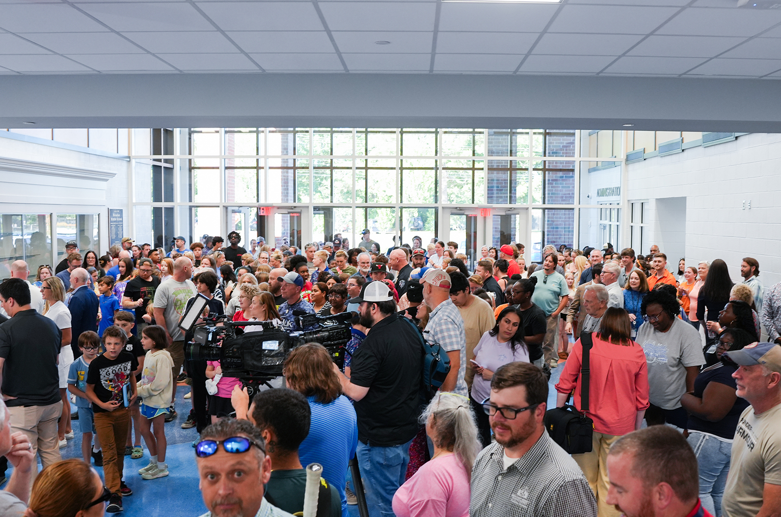 A large group of people gather indoors, some wearing glasses, hats, and sunglasses. A camera crew films the scene.