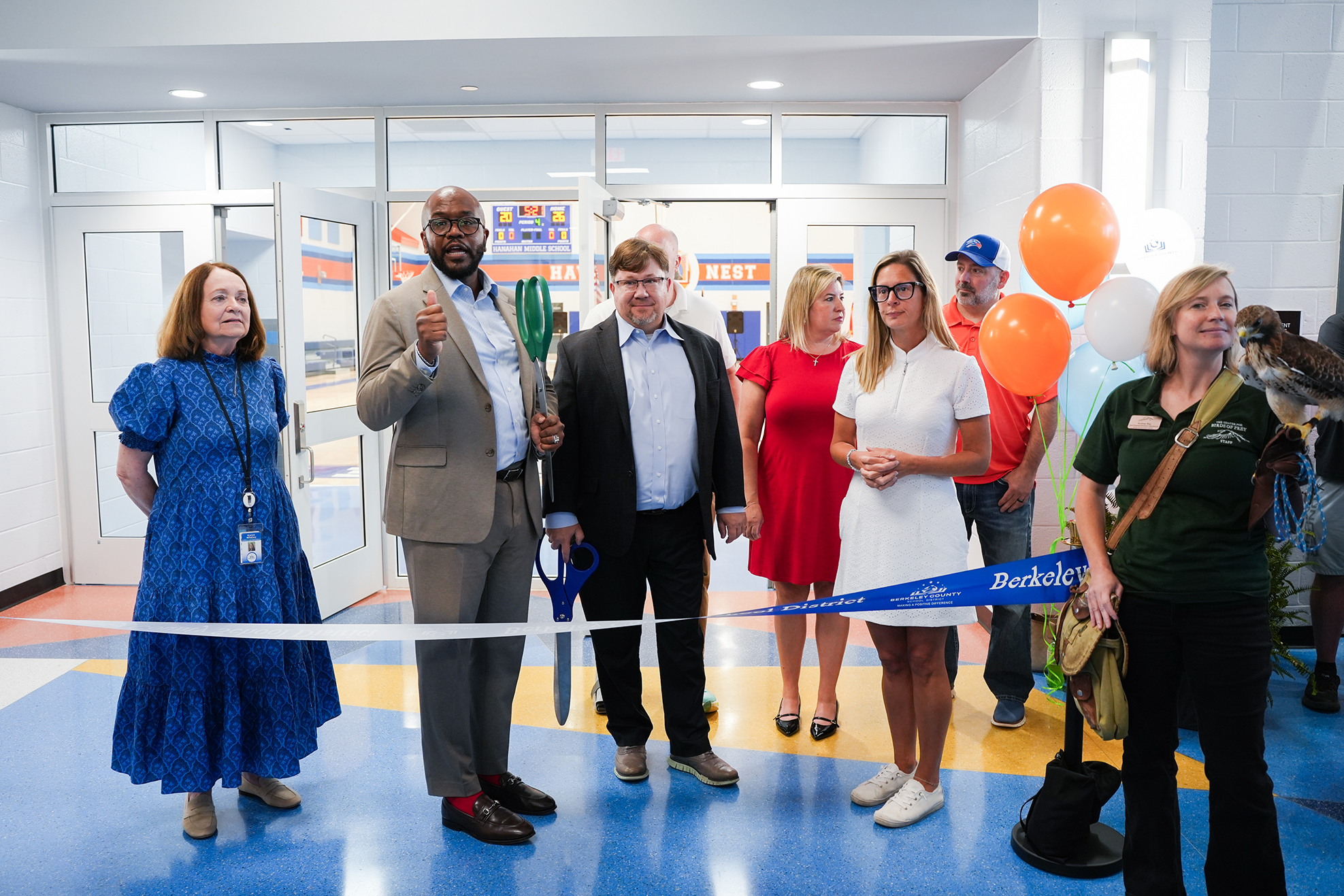 A group of people stand in a hallway holding a ribbon, ready to cut it. Colorful balloons are present.