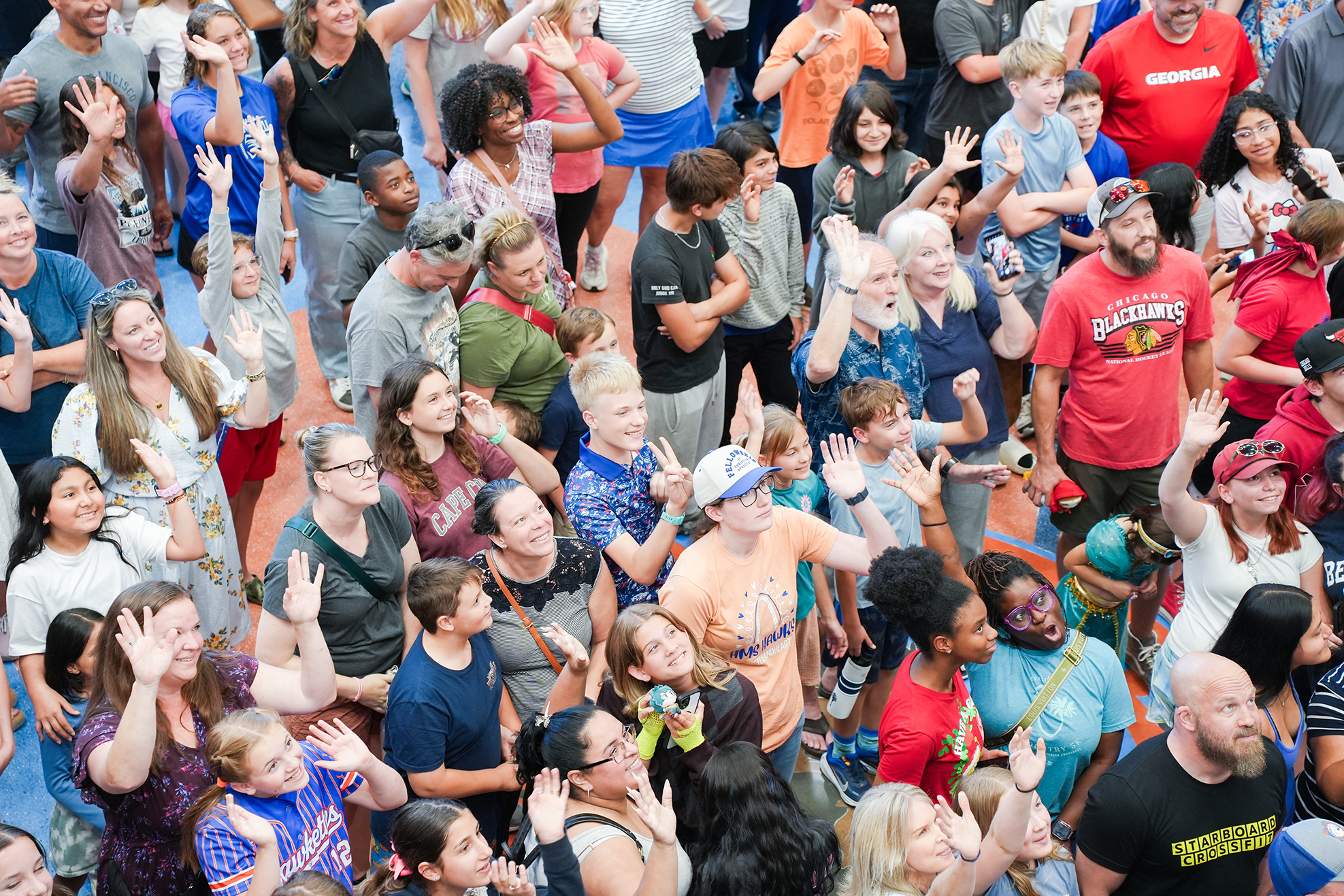 A crowd of people in various clothing and colors, many raising hands, standing and interacting.