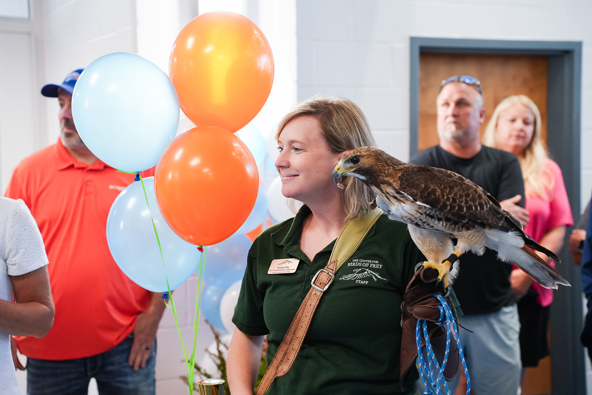 A woman holding a bird of prey on her arm stands among a crowd. Colorful balloons are in the background.