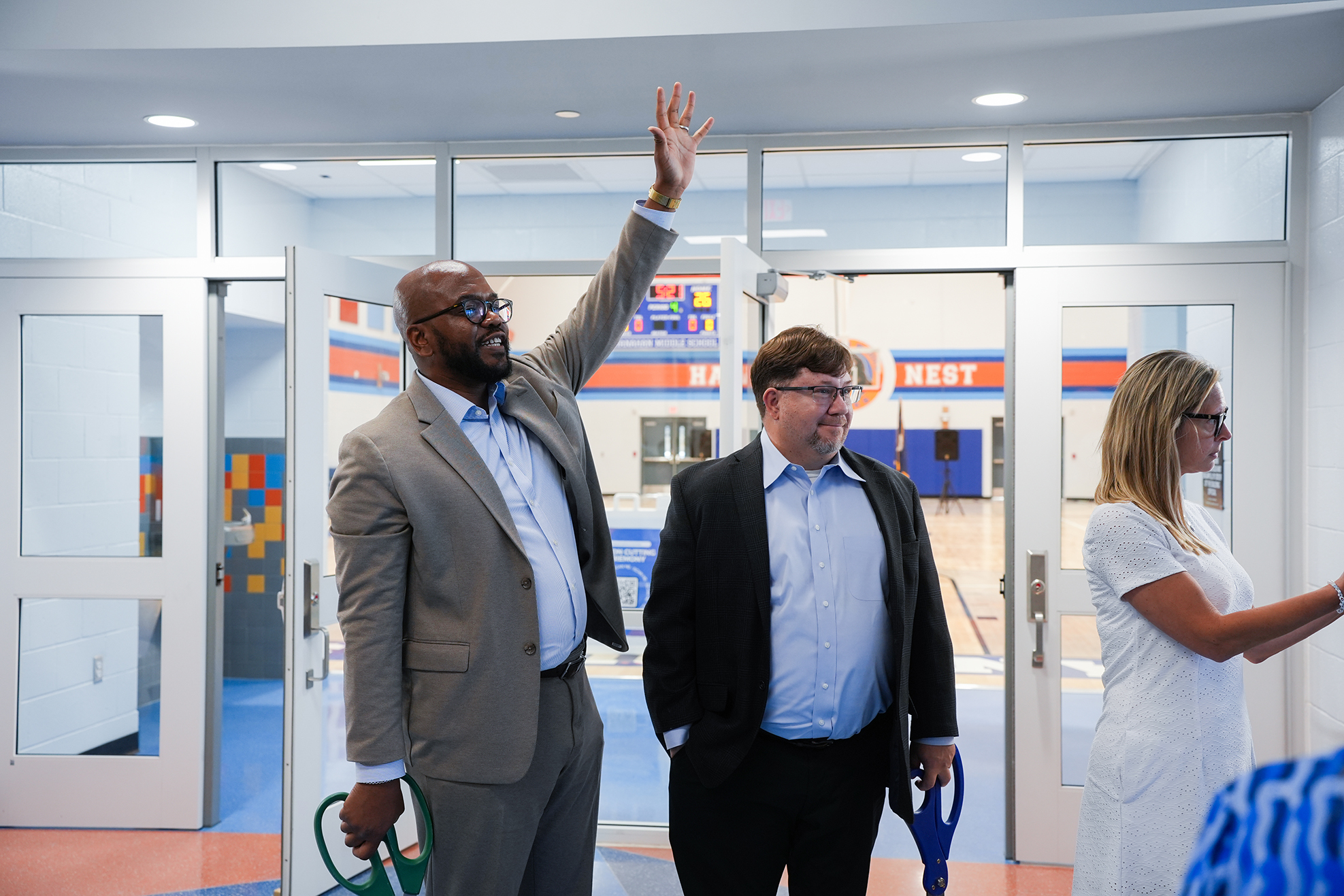 Three people stand inside a room with white walls. Two men and a woman face the camera. One man waves.
