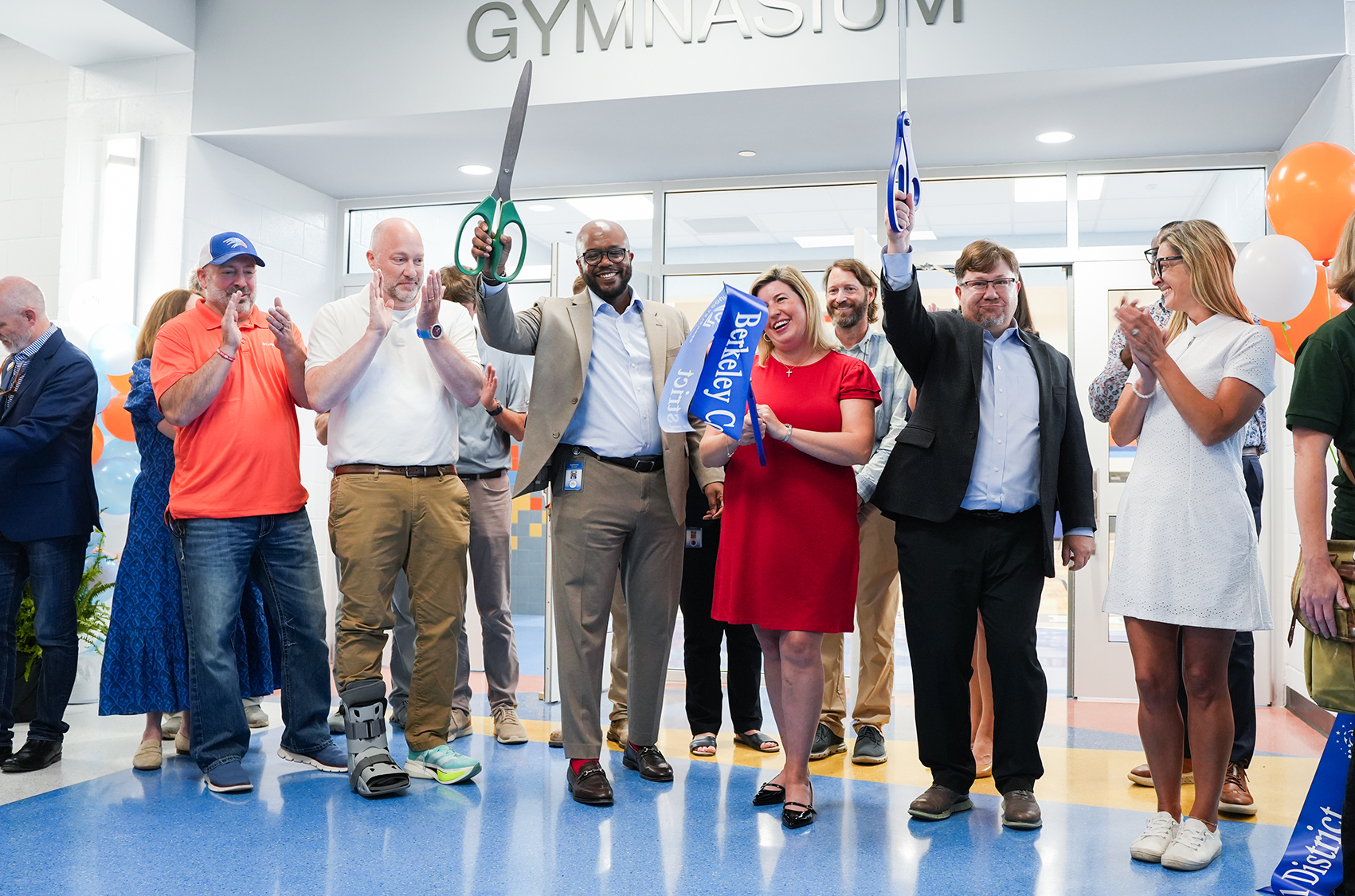 A group of adults stand in a brightly lit hallway, one man holds scissors and another holds a blue ribbon.