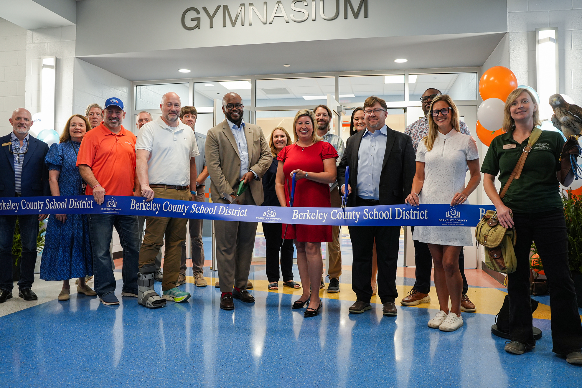A group of people stands in front of a ribbon for an event at the Berkeley County Schools gymnasium.