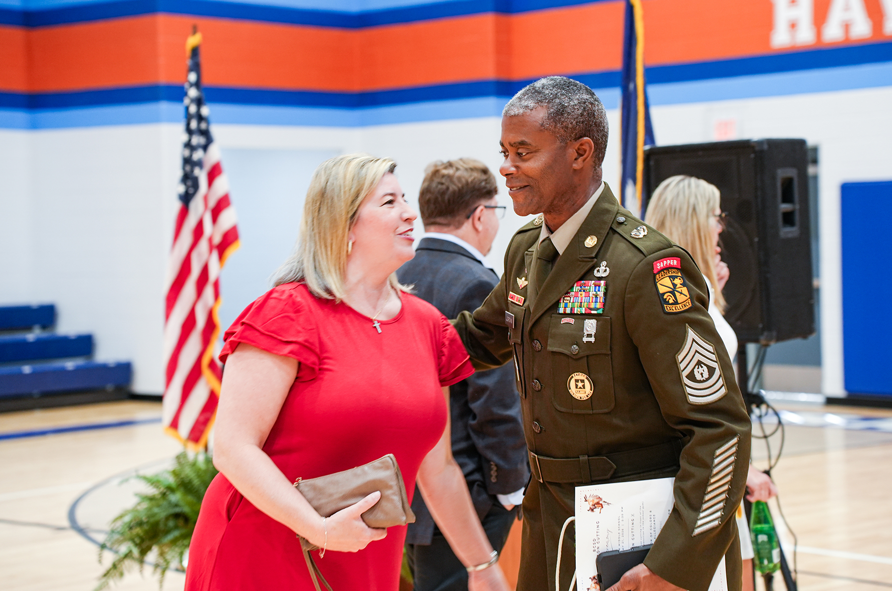 A man in a military uniform talks to a woman in a red dress inside a gym.