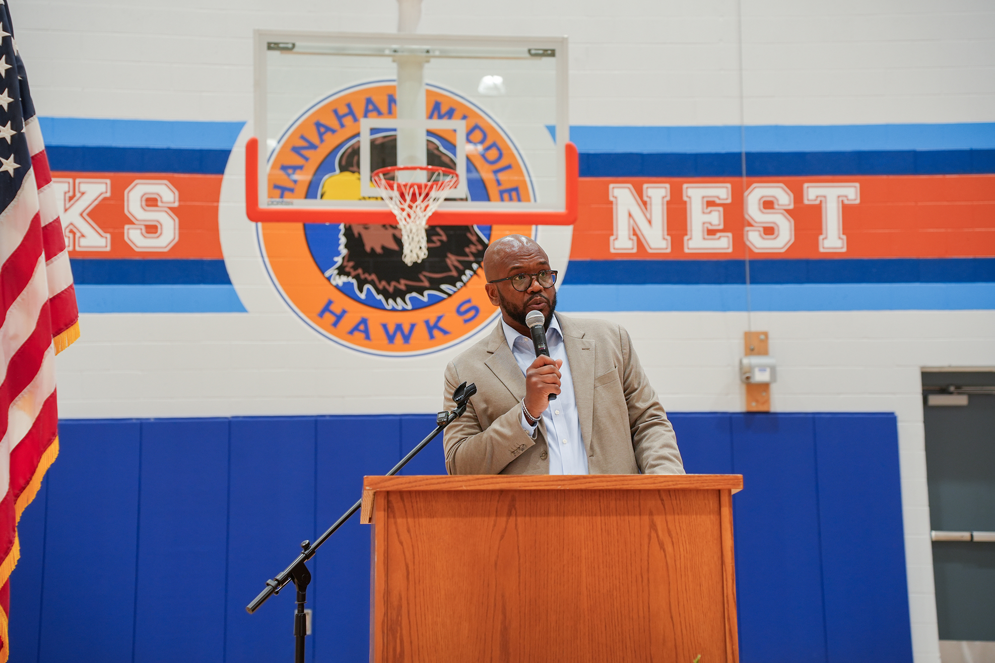 A man speaks at a podium with a microphone, in front of a wall with a basketball logo.