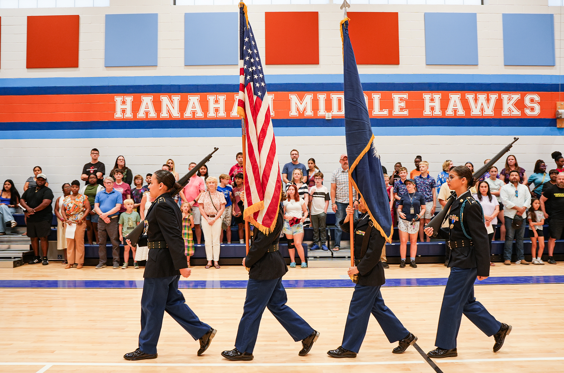 Four individuals in uniform walk across a basketball court, carrying flags. Spectators watch from bleachers.