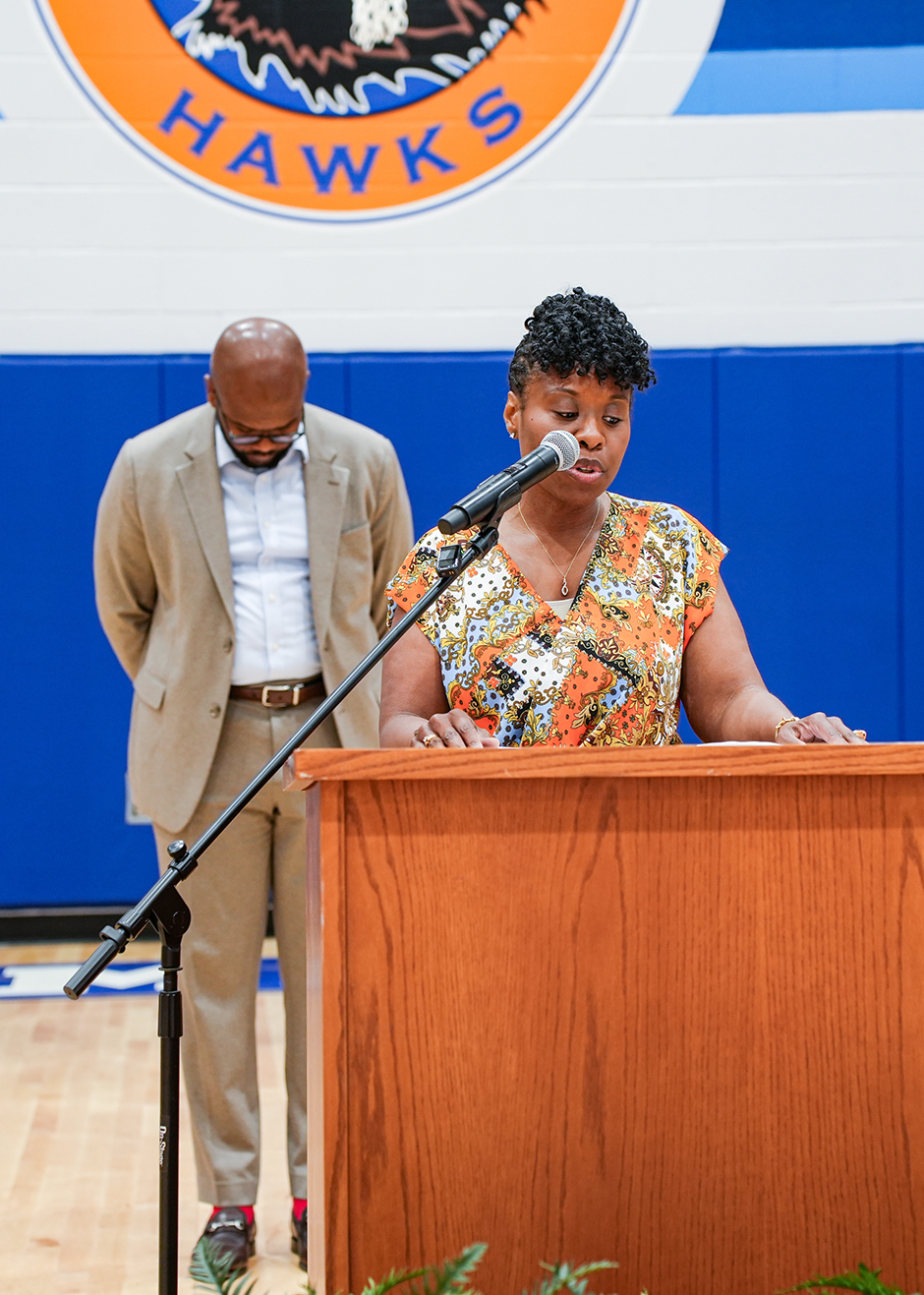 A woman stands behind a podium and speaks into a microphone, with a man standing behind her, both in a gym.