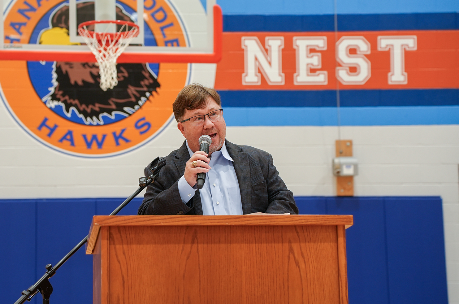 Man in glasses speaks into microphone at a podium in front of a Hawks logo and a basketball hoop.