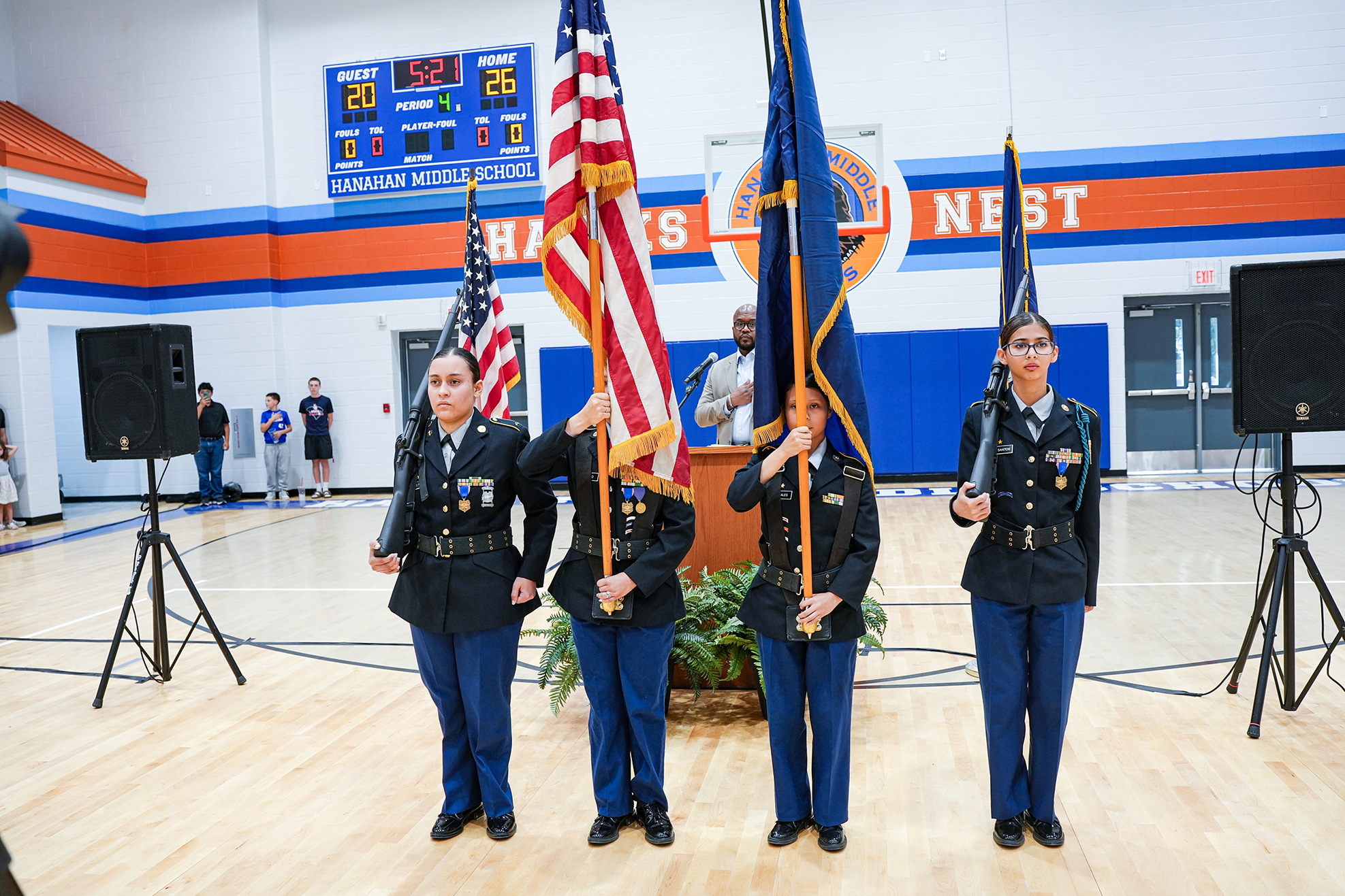 Four women in uniform stand in a gymnasium, holding flags. Two flags are raised while two are lowered.