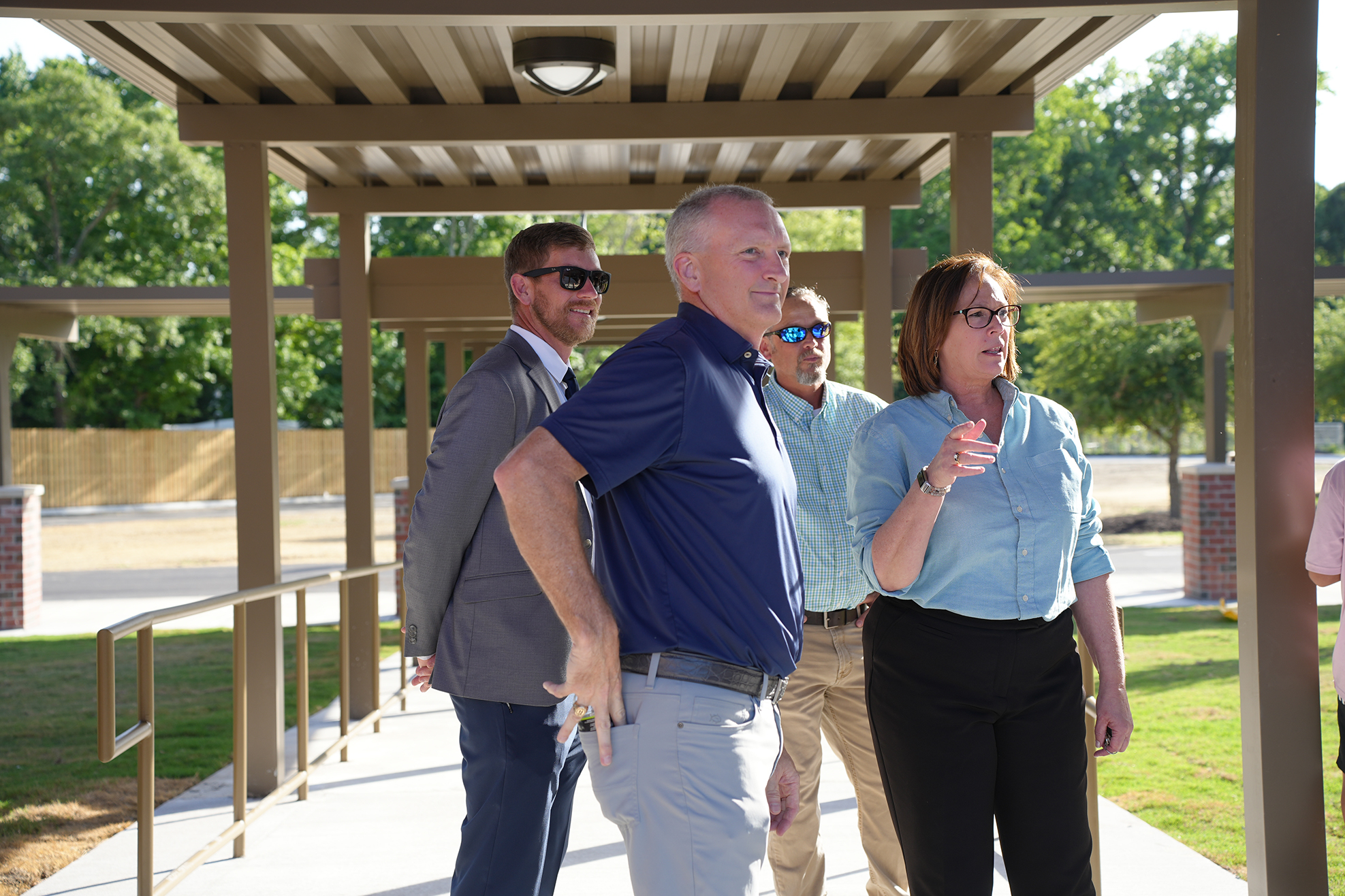 Four adults stand on a covered walkway. Three are facing forward. A brick wall is behind them.