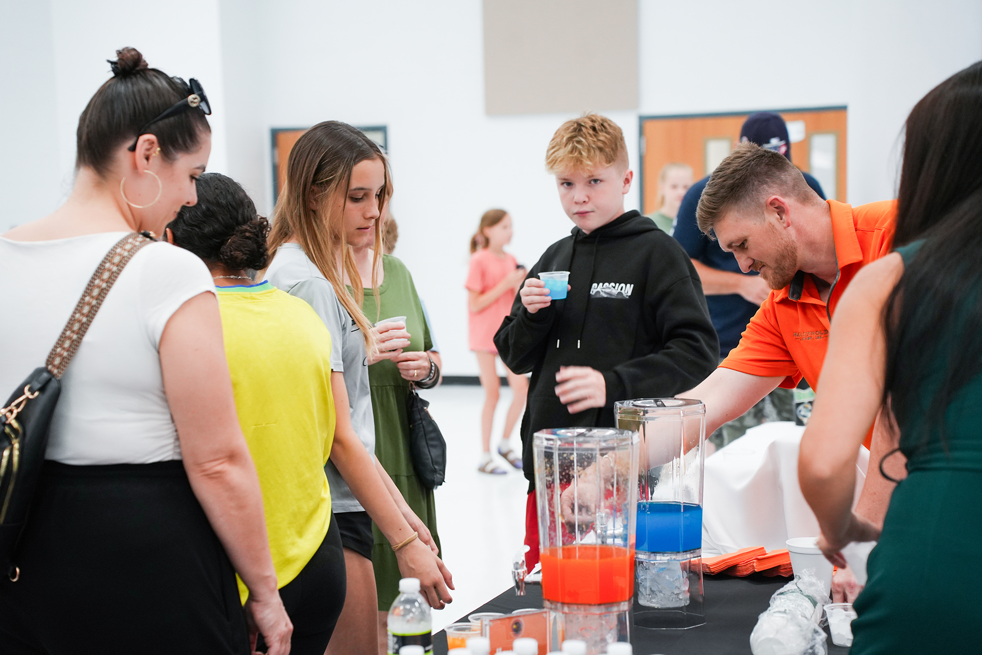 A group of adults and children gather around a table, examining items. One person pours a liquid.