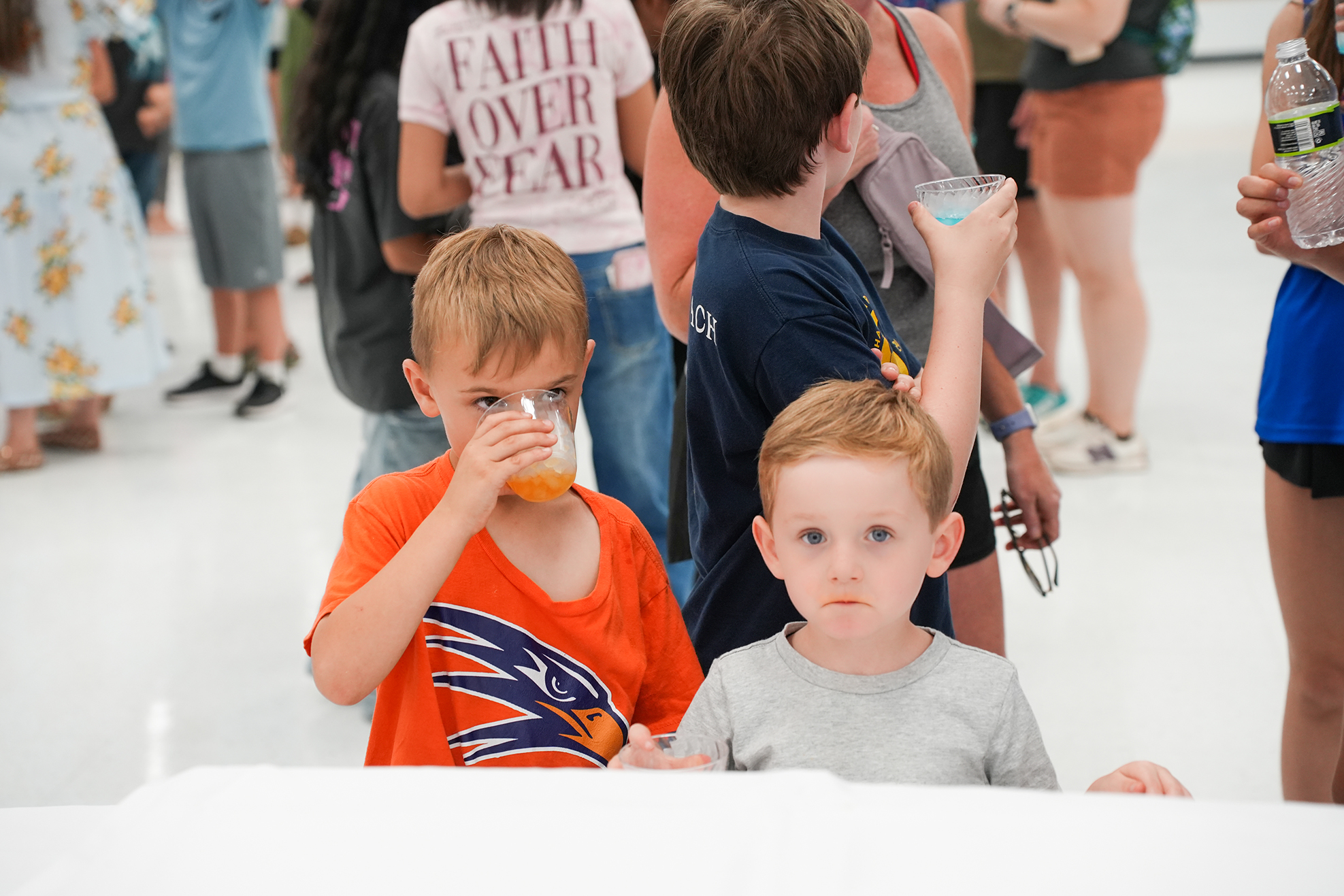 Two young boys drink beverages. One wears an orange shirt, and the other wears a gray shirt. Several people stand in the background.
