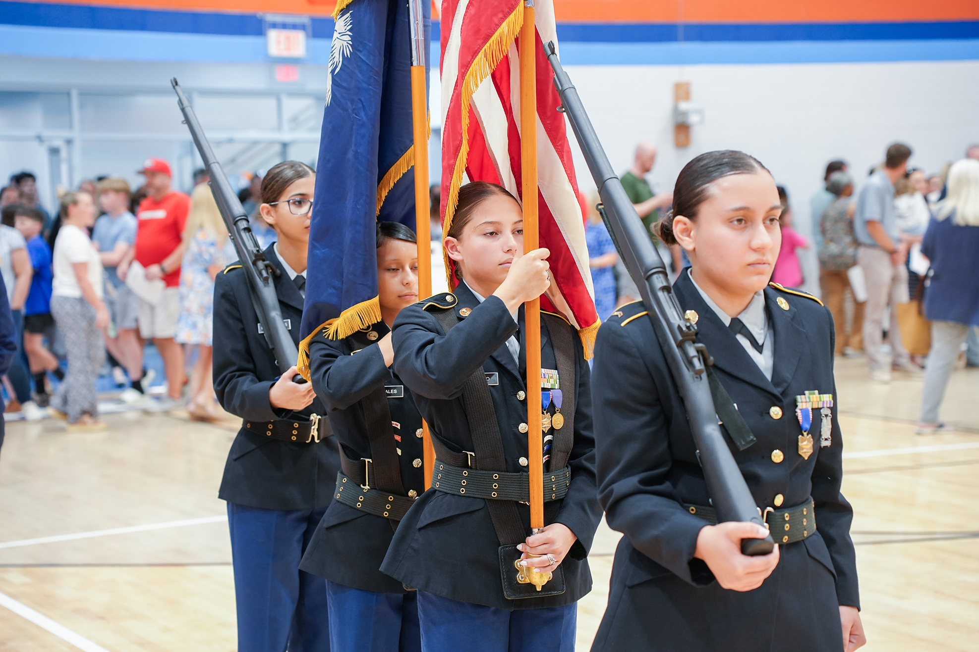 Four women in uniform hold flags in a gymnasium. Behind them, a crowd stands on the sidelines.