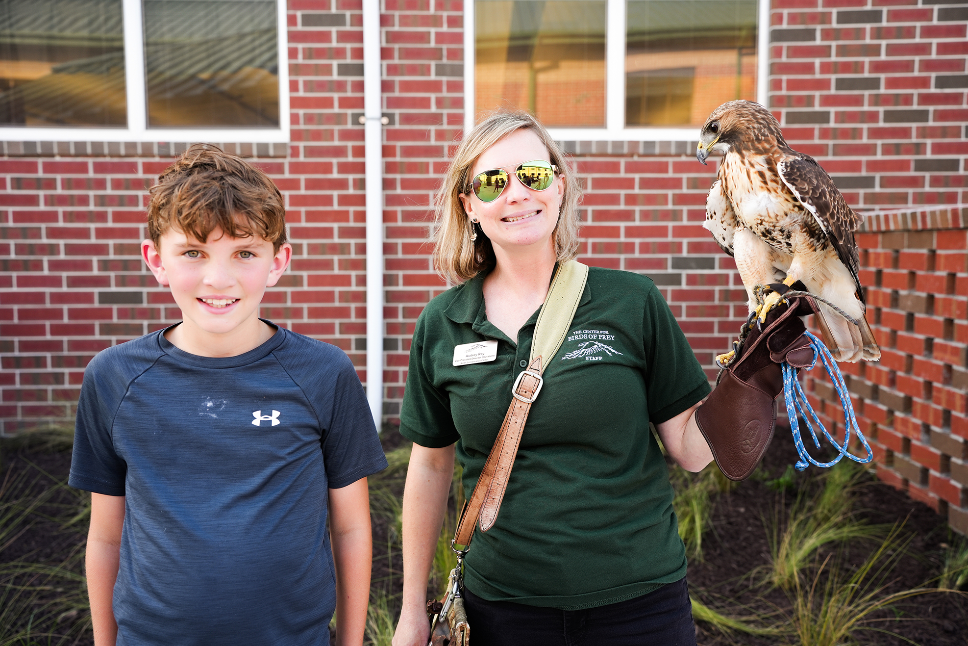 A woman in green holds a hawk on her arm, next to a smiling boy in blue. They stand before a red brick building.