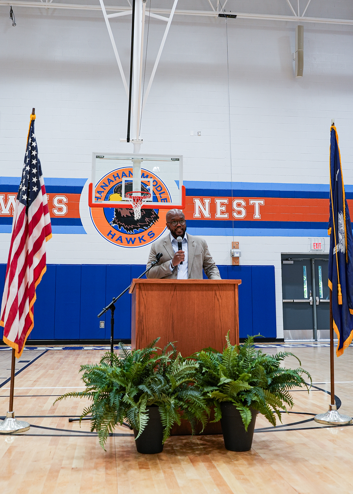 A man speaks at a podium with American flags and plants in front, inside a gymnasium with a basketball court.