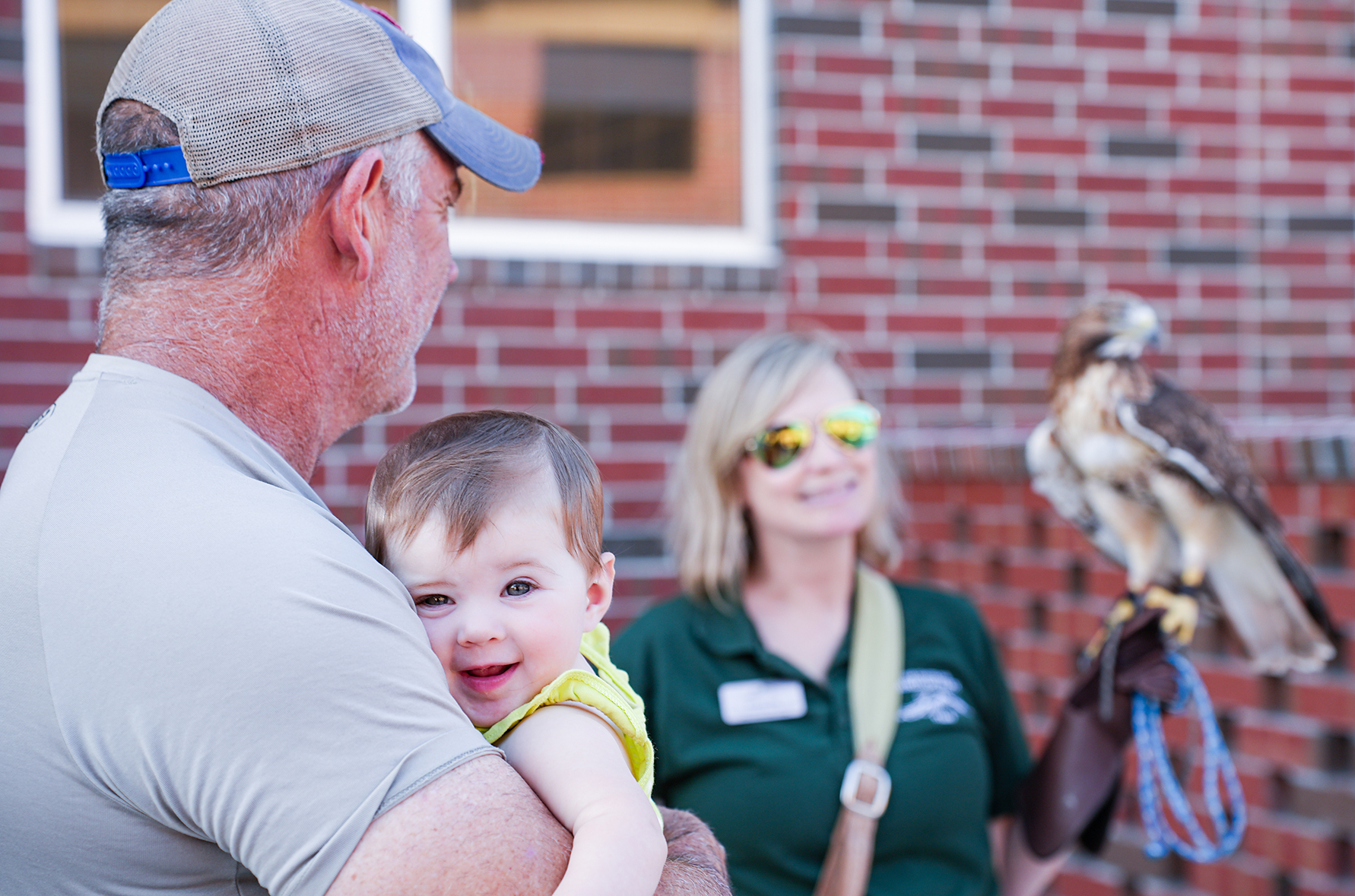 A man holding a baby smiles at a woman holding a falcon. Brick wall in background.