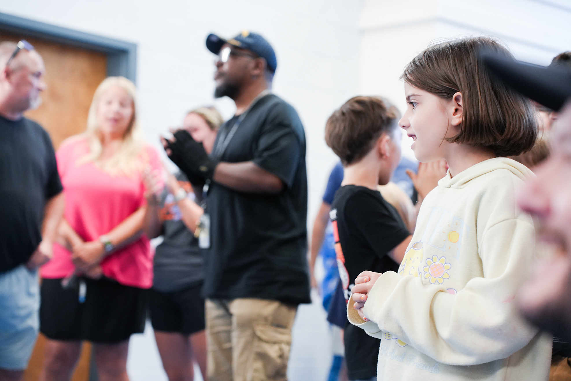 A group of people standing indoors. A girl in a white sweatshirt is looking to the side.