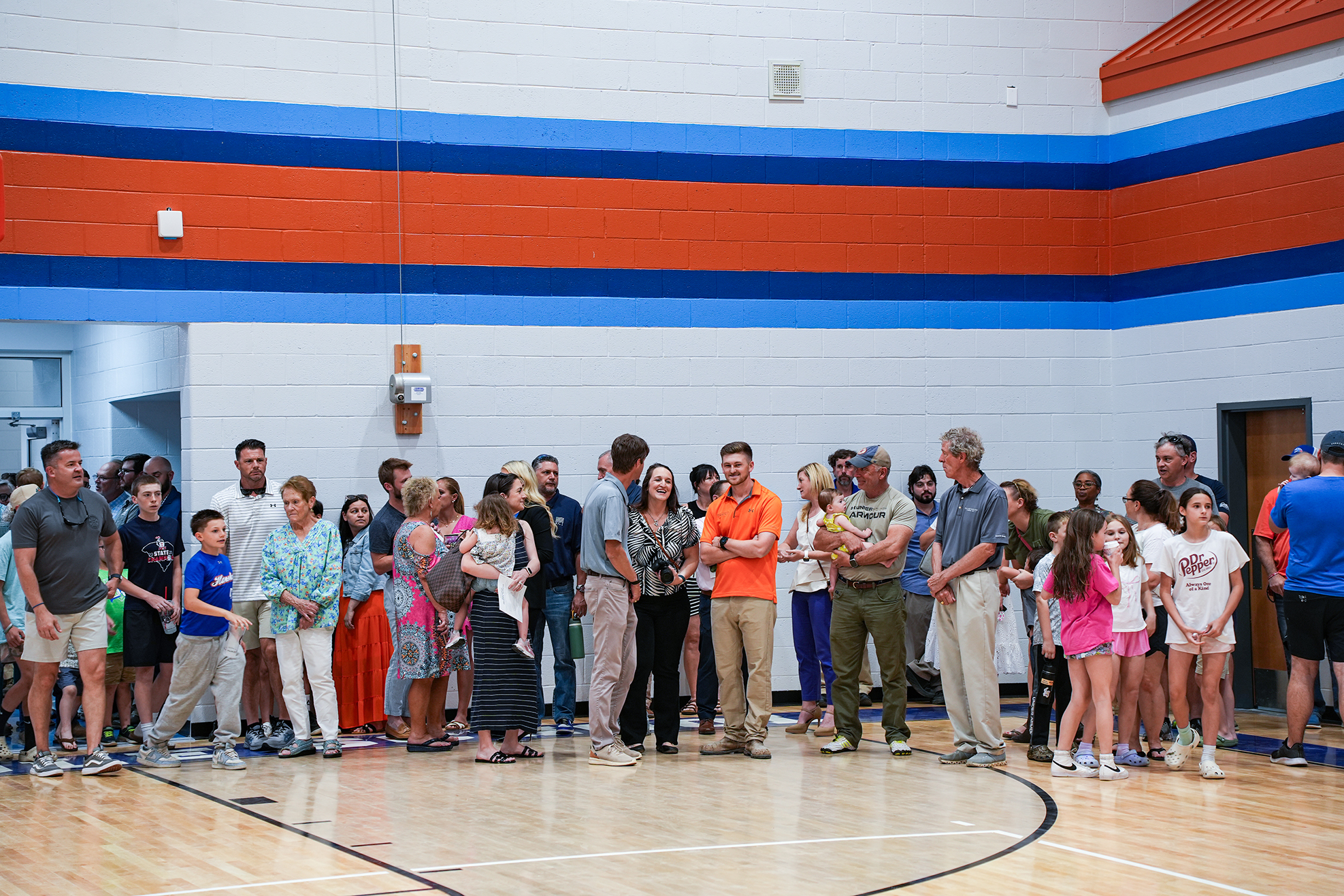 A crowd of people gathers in a gym, some wearing casual attire and shoes, on a wooden basketball court.