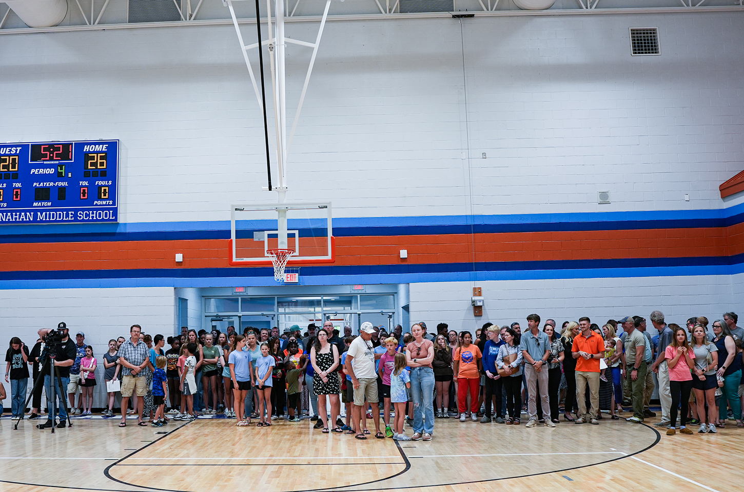 A gym with a basketball court and hoop; many people stand, some with cameras, and a blue scoreboard is on the wall.