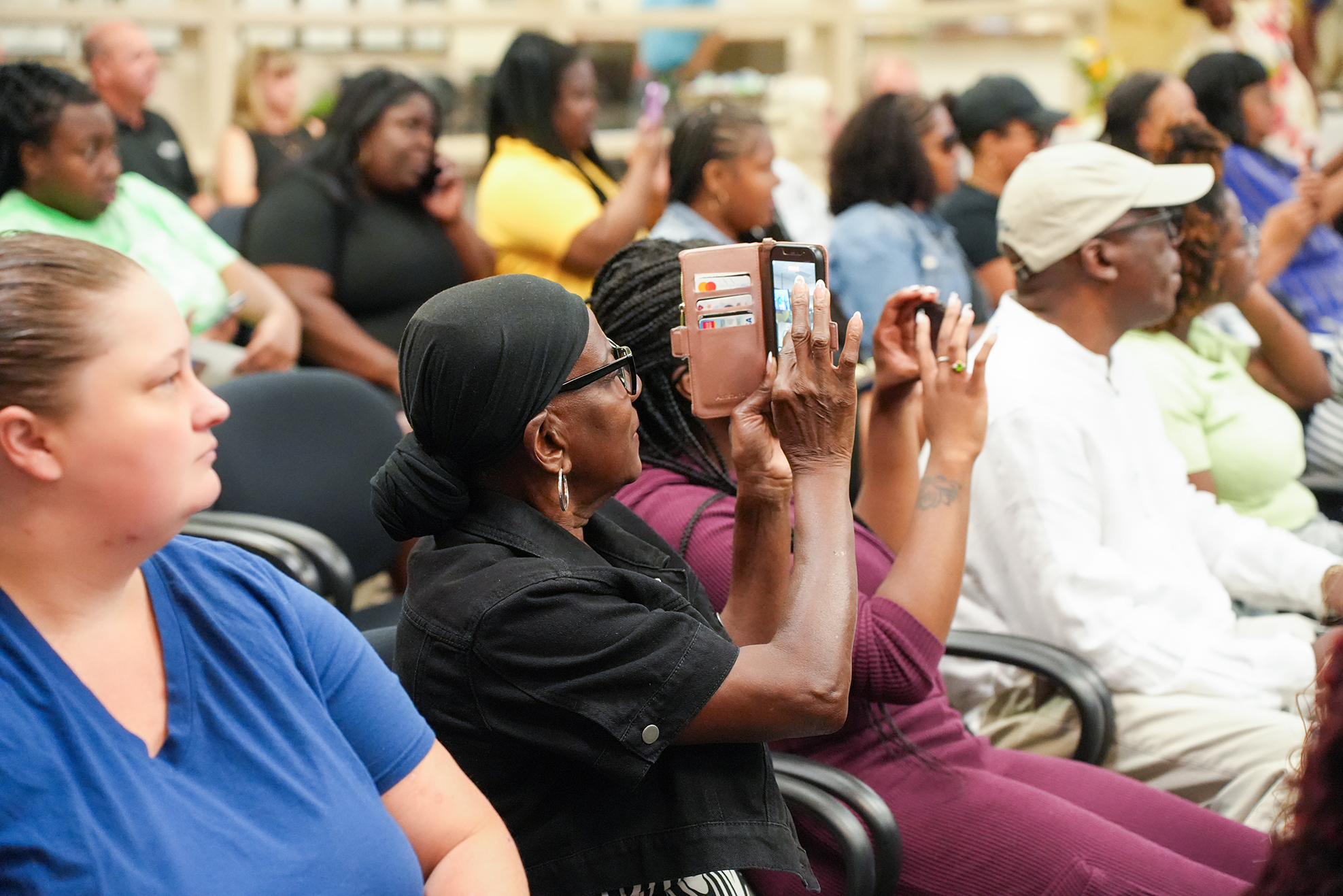 Several people in a meeting, some with phones, sitting on chairs, one person in a white hat.