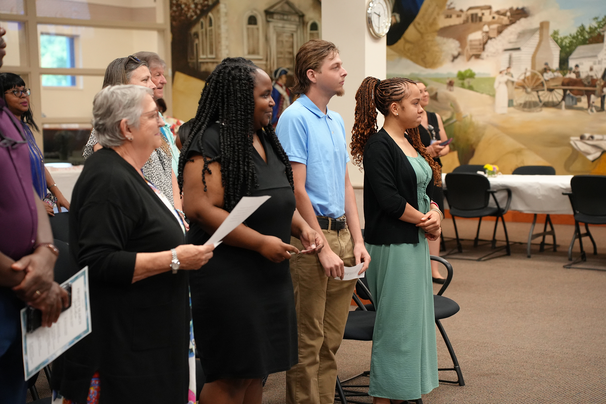 Group of people stand together in a room with a mural behind them, holding papers.