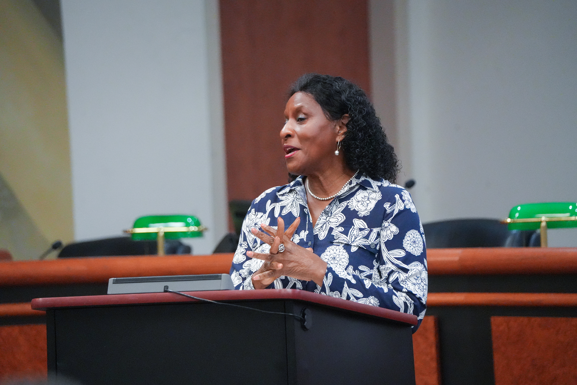 Person in blue floral blouse speaking at a podium, with chairs, desk, and green desk lamps behind.