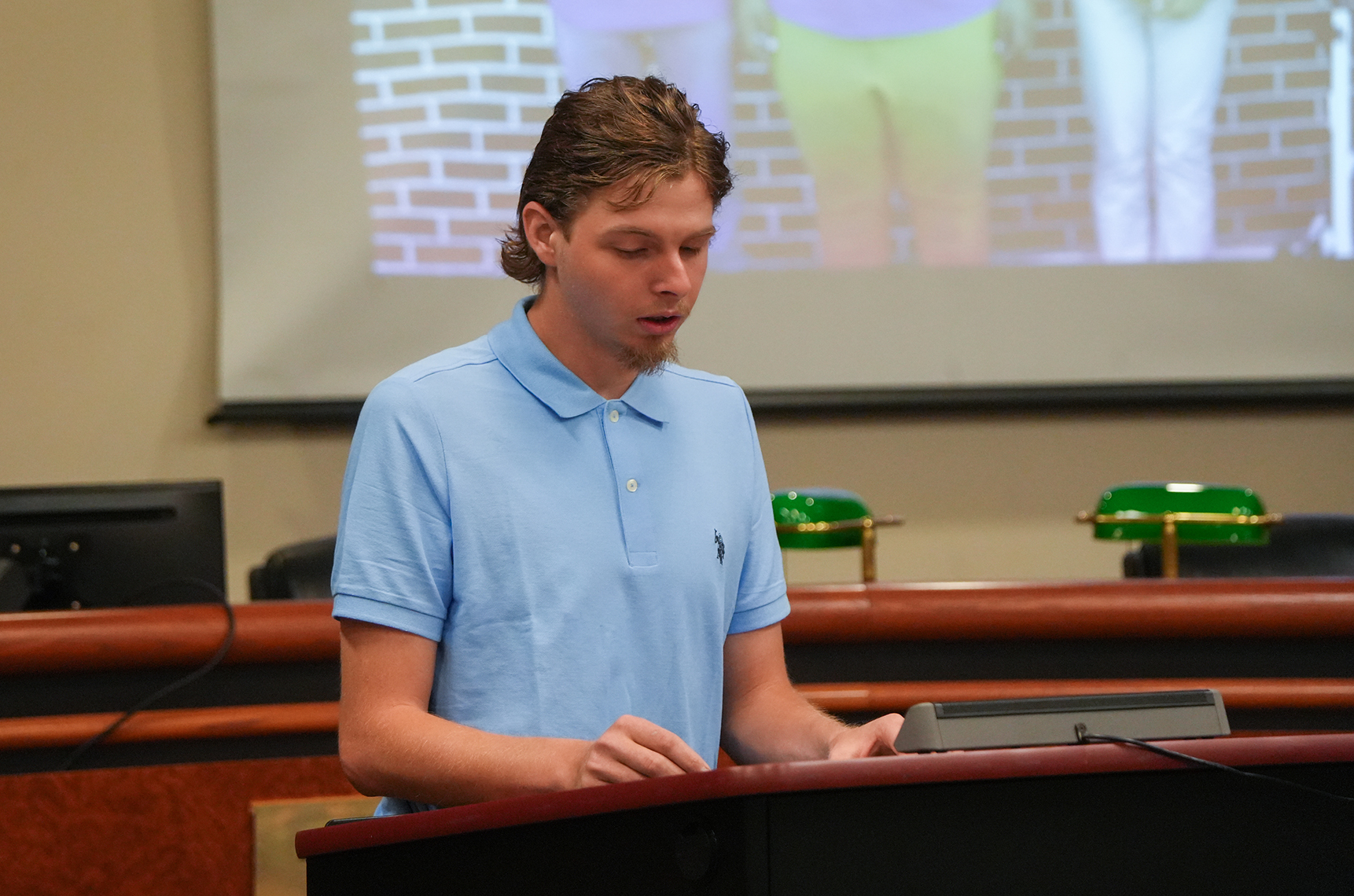 A man stands at a podium with a projector screen behind him, showing a brick wall and legs.