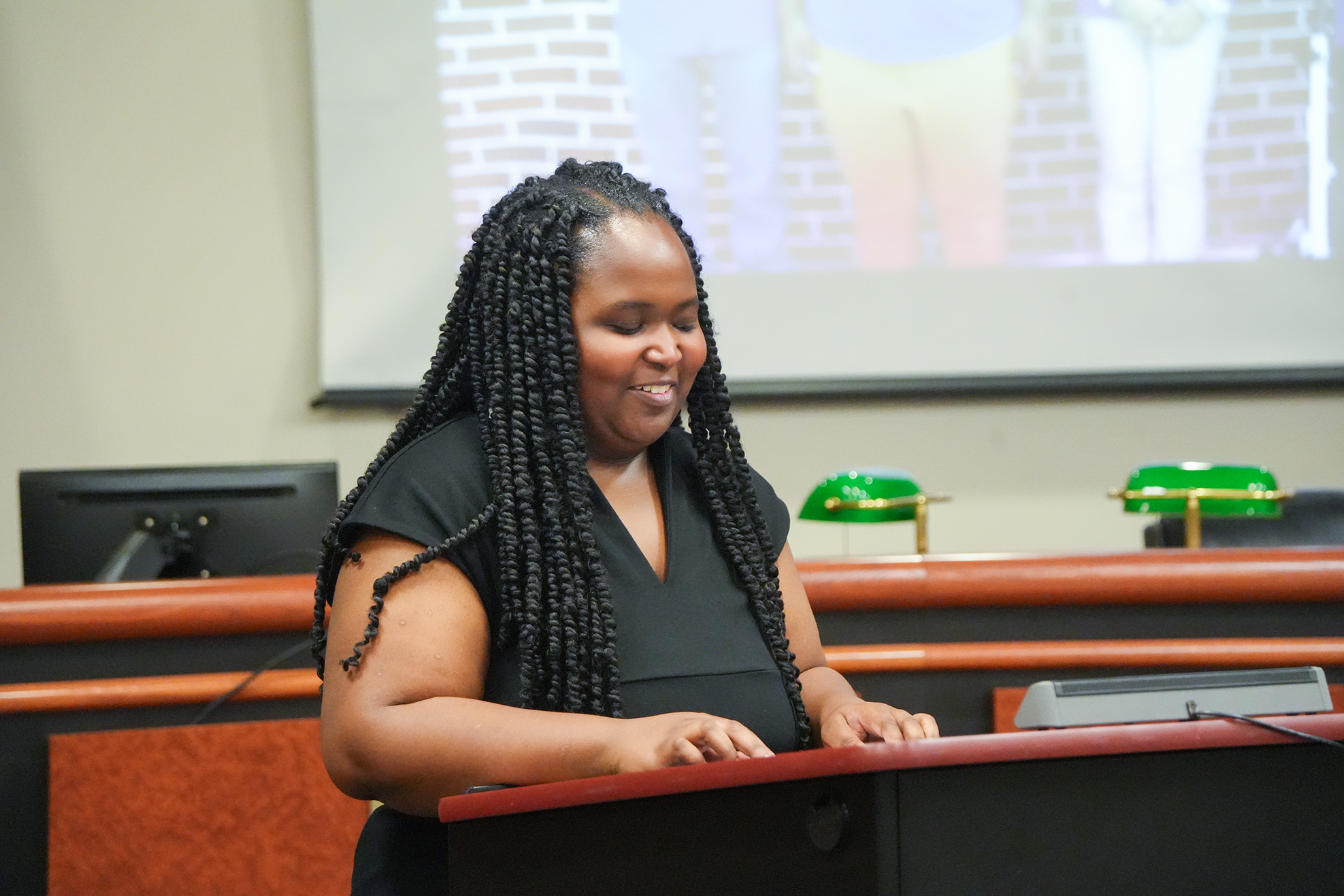 Woman standing at a desk with hands on top, smiling. Behind her, a projector screen displays an image.