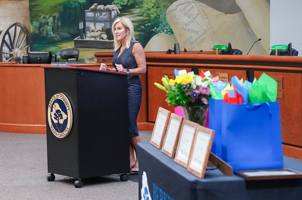 A woman stands at a podium, speaking with a microphone. Behind her, a wall mural features a landscape.