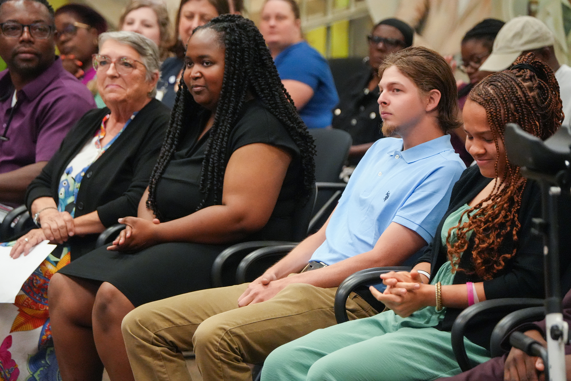 People sit in rows of chairs; a woman in a black dress speaks and a man next to her listens.