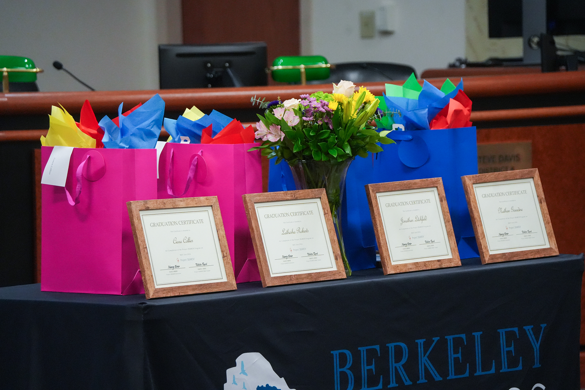 Four diplomas and gift bags on a Berkeley banner-covered table. The table is in a room with a podium.