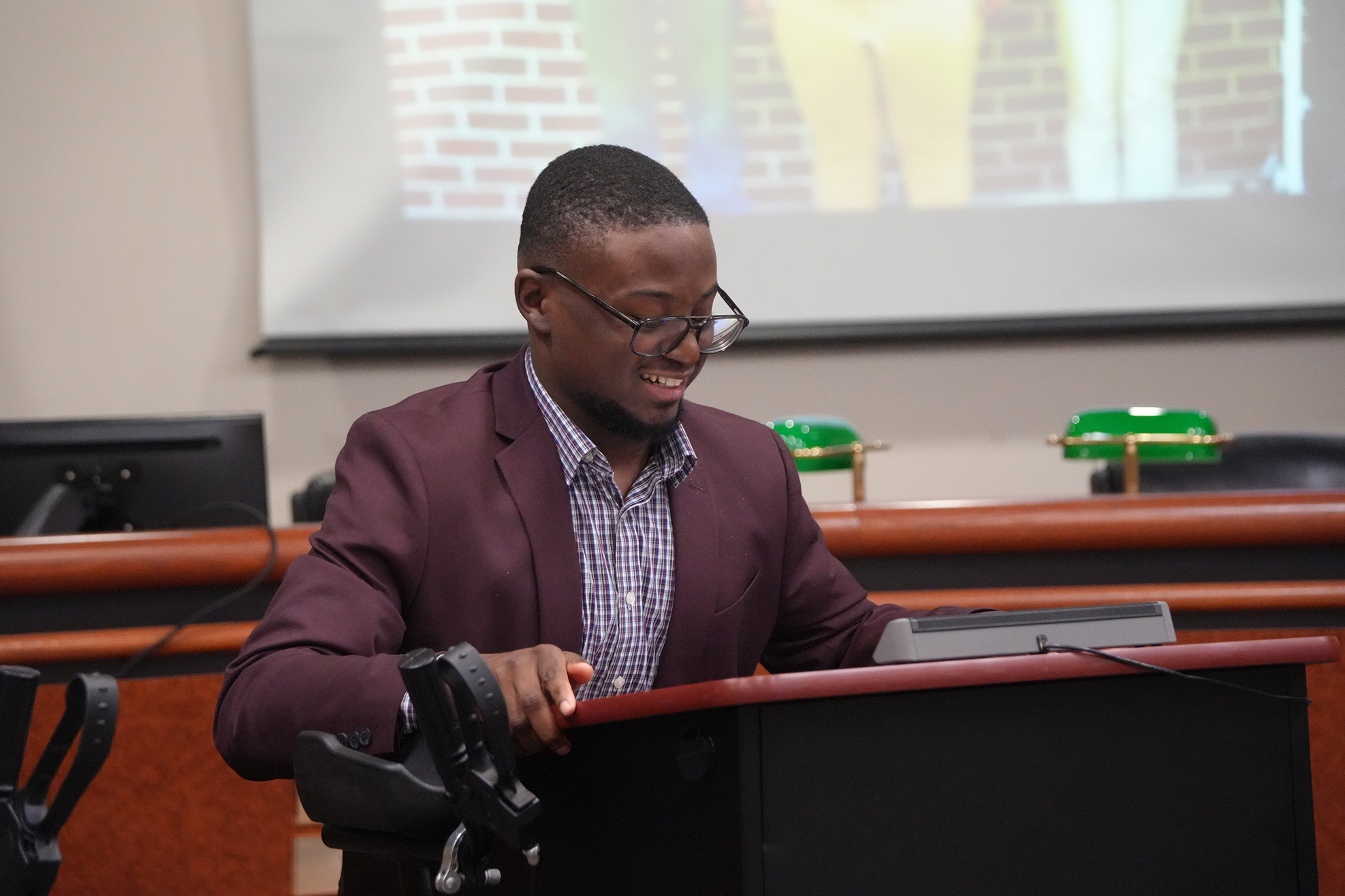 A man wearing glasses sits at a desk, speaking while a projector screen displays a brick wall.