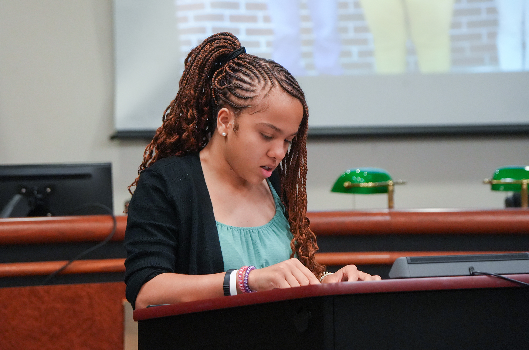 A young person with curly hair and a black jacket gives a presentation at a podium.