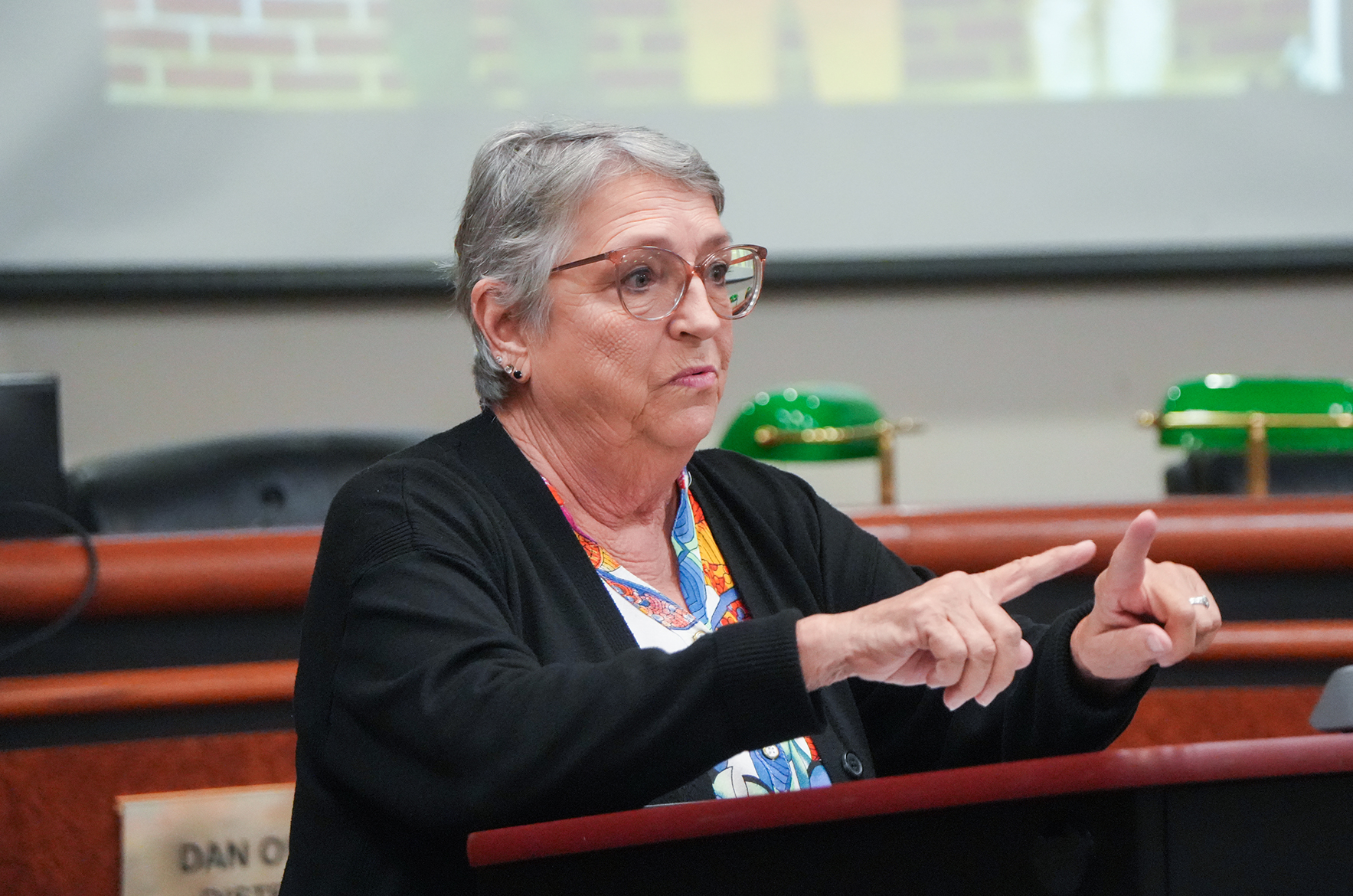 Older woman with glasses seated at a podium speaking and gesturing with her hands.