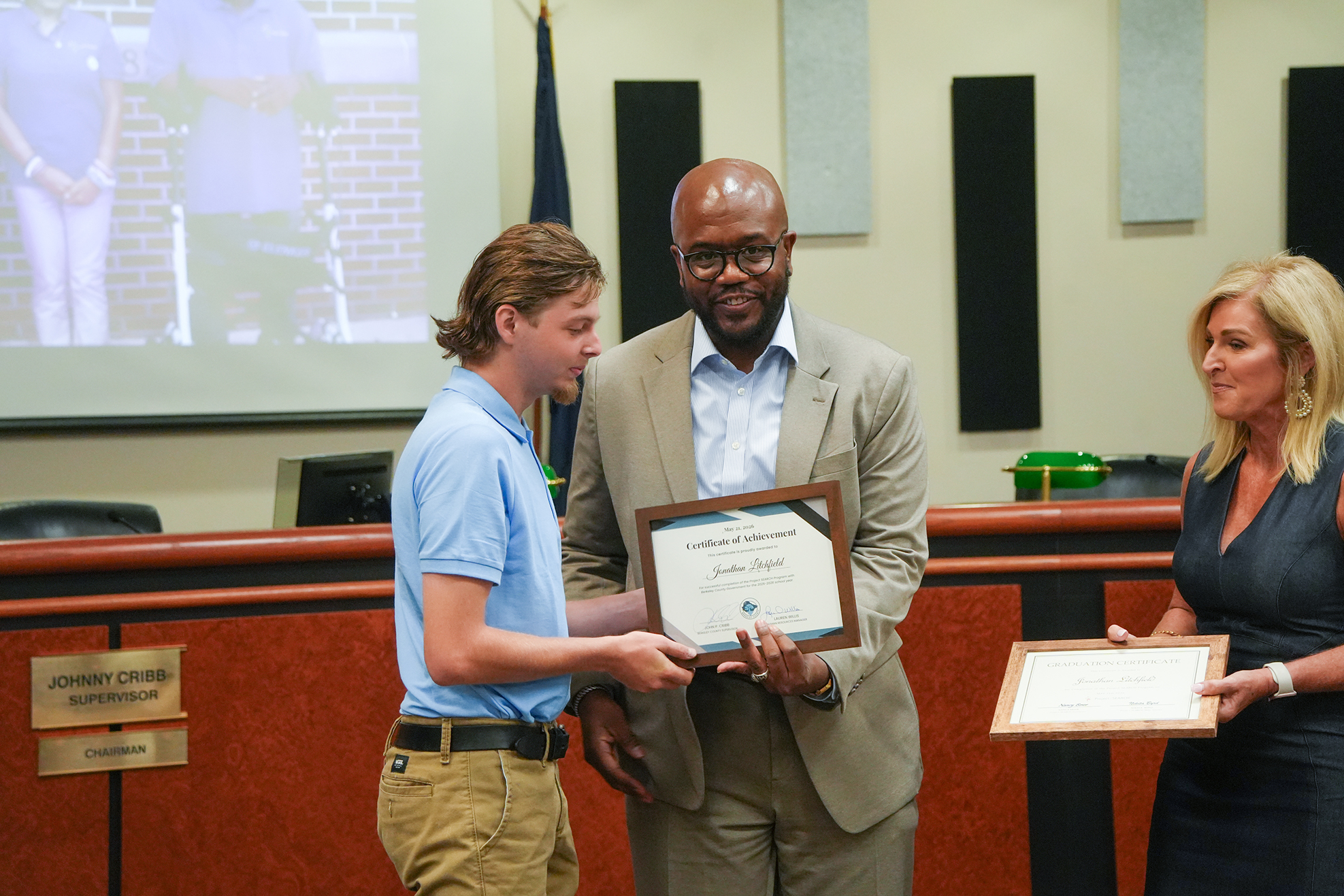 A man in a suit presenting a framed certificate to a young man in a blue shirt.