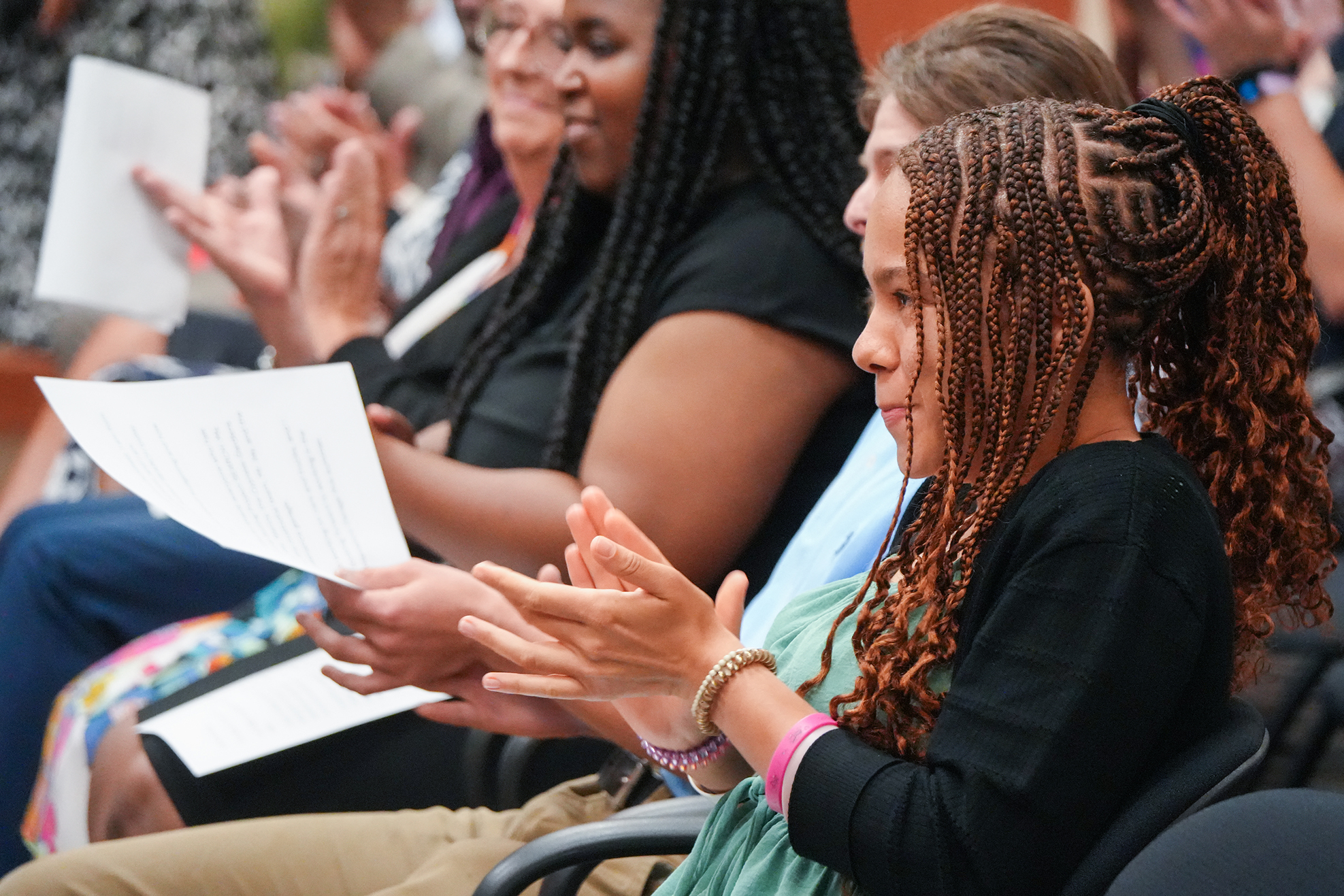 People in a room, seated and clapping. One person holds a paper and claps. Braided hair visible.