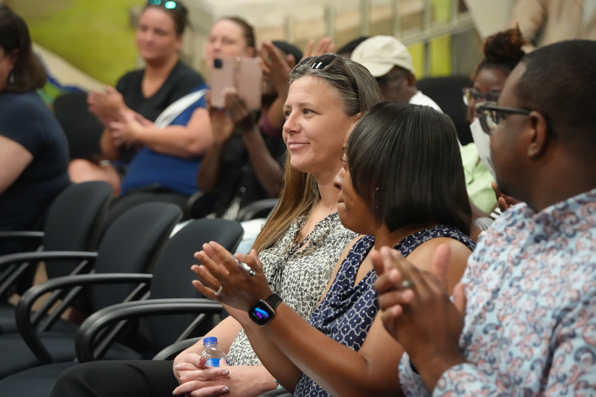 Several adults sit in chairs in an auditorium, clapping. Some have masks on.