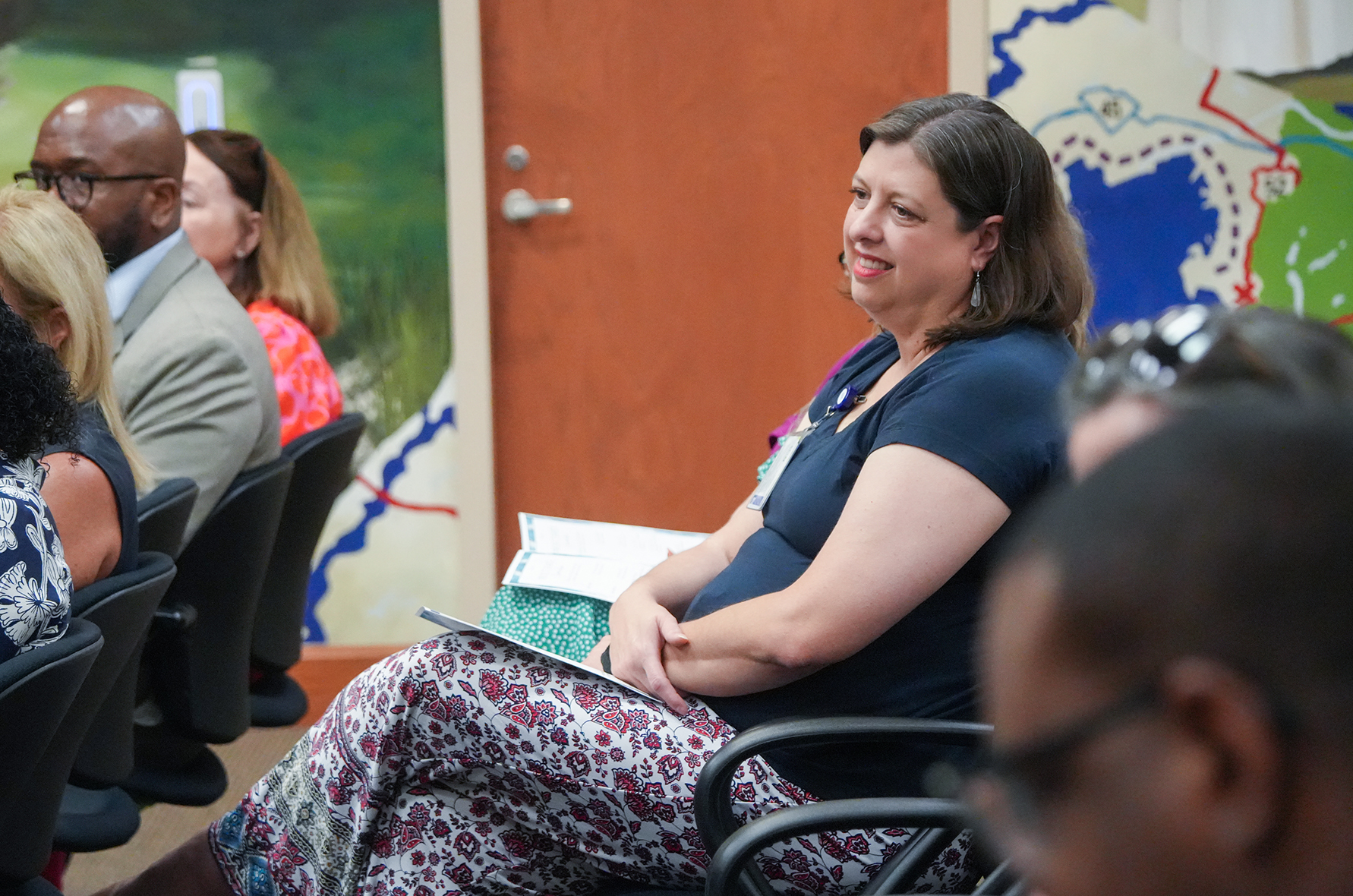 A woman sits among others in a conference room, smiling and holding a book. Behind her, a map adorns the wall.