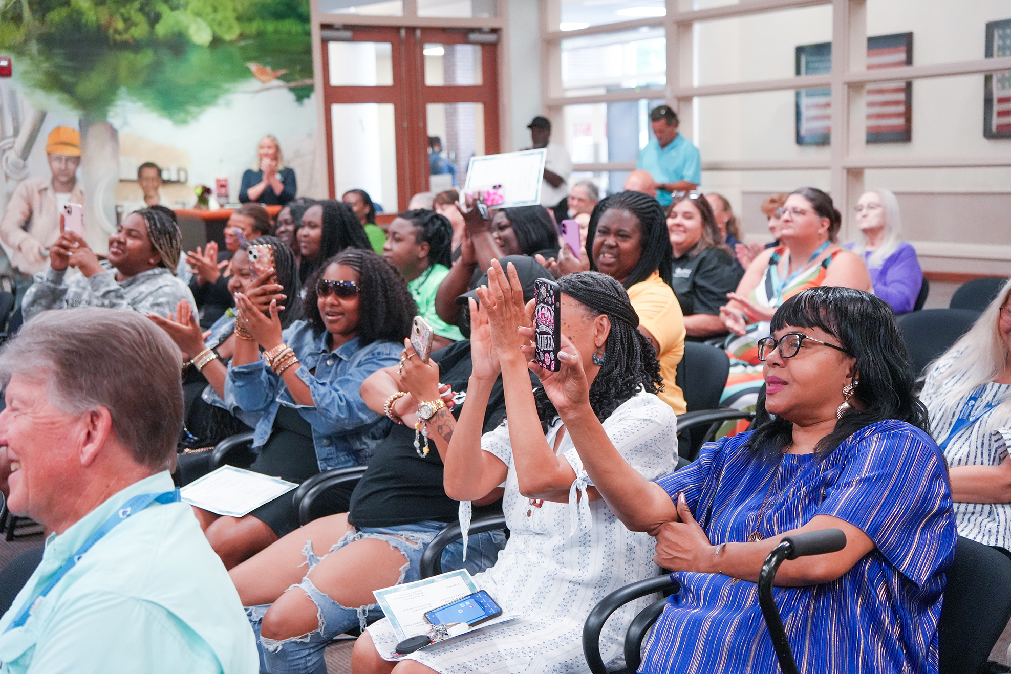 Many people, seated in chairs, are clapping and smiling, with a few using their phones. Background includes a mural.