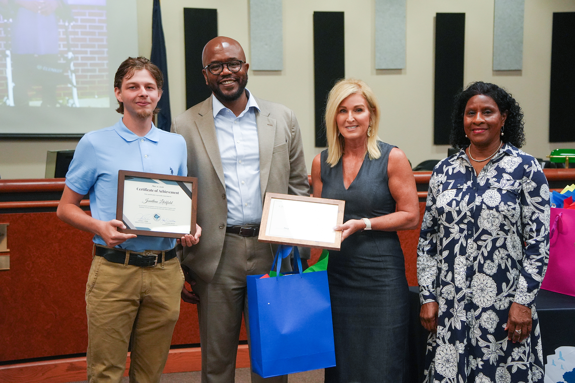 Four people stand together, holding awards and a blue bag. A man and woman hold certificates, and a woman holds a bag.