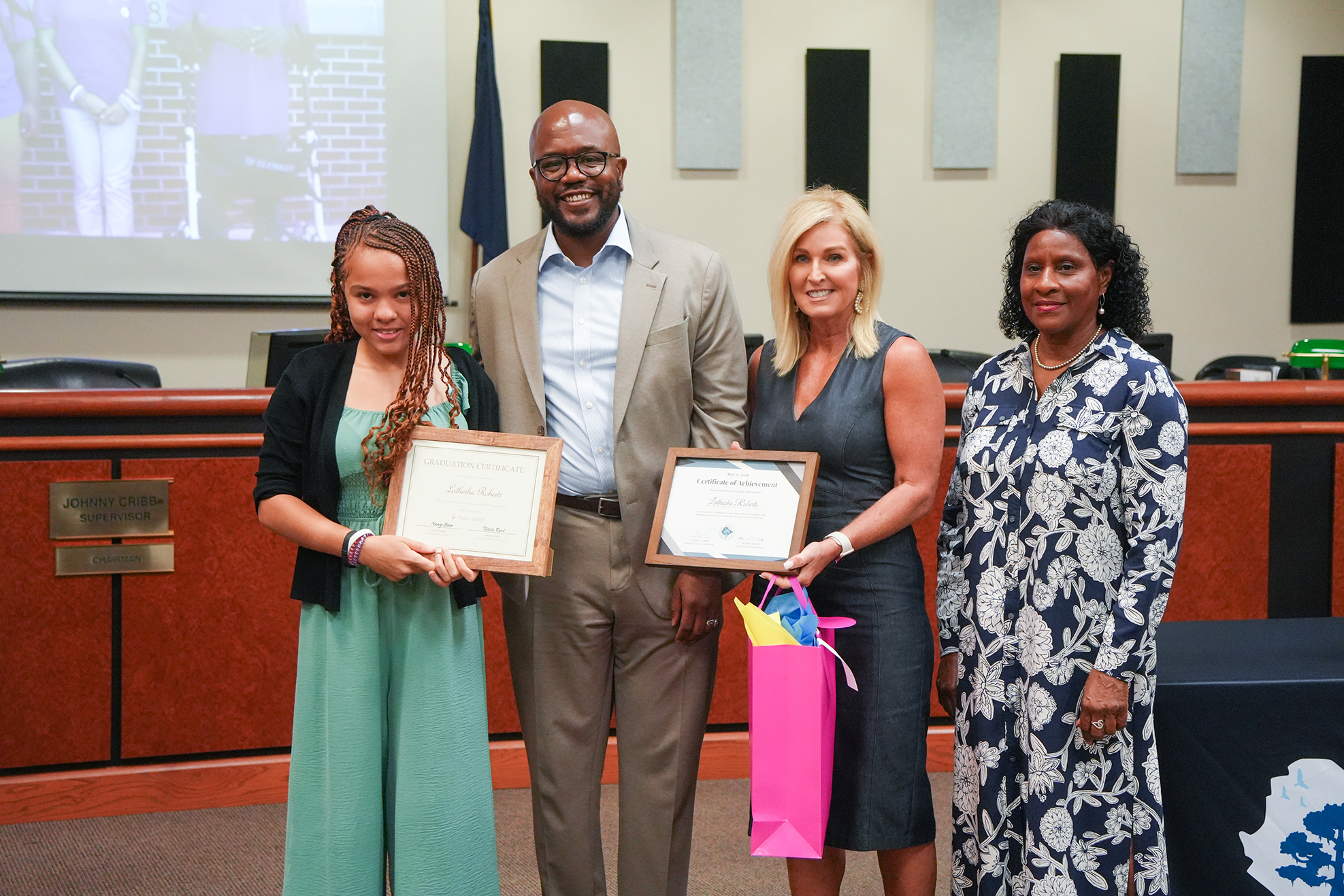 Four adults and a young girl hold certificates in front of a podium in an indoor room.