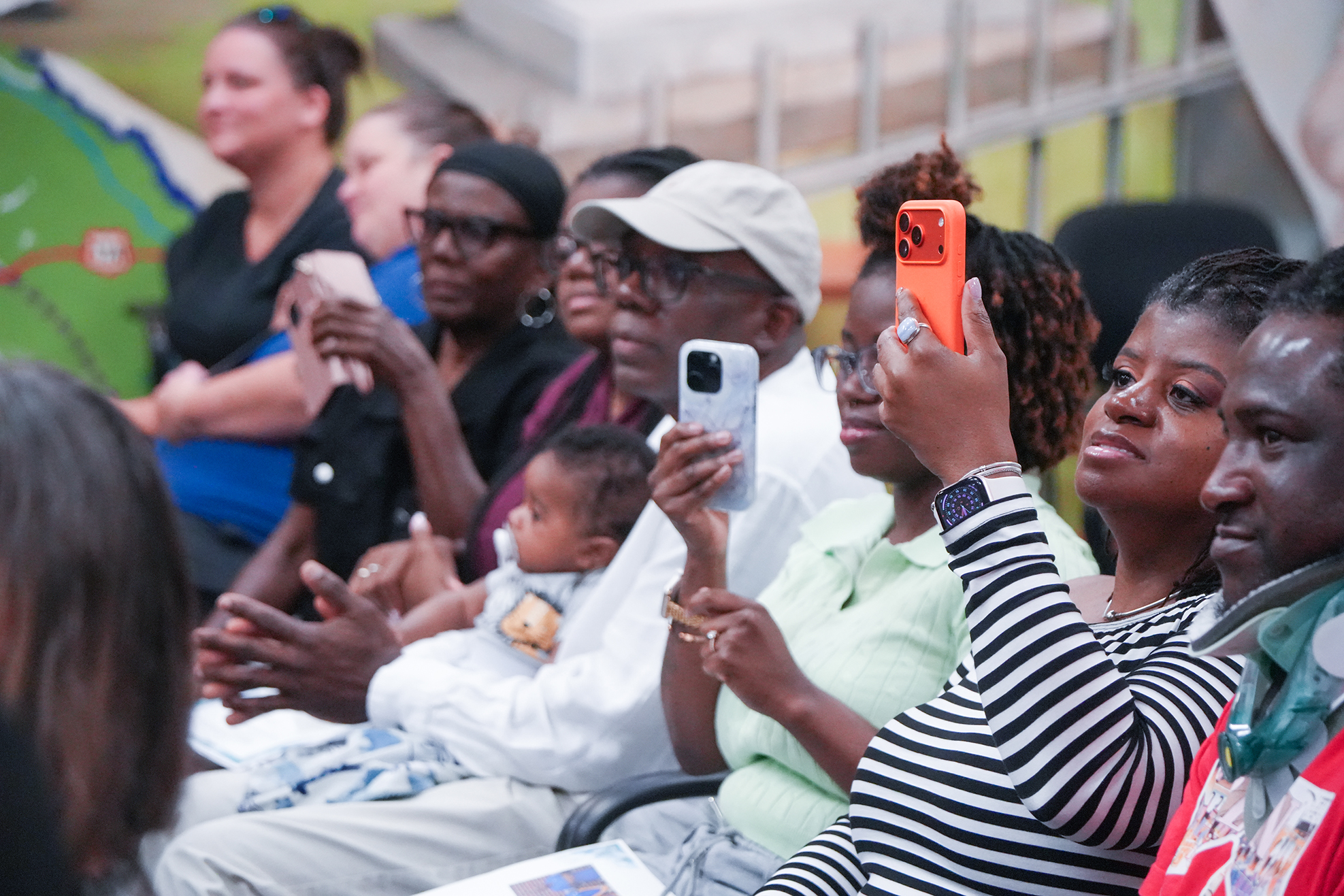 A group of adults and a baby seated in a room, some with phones held up, clapping.