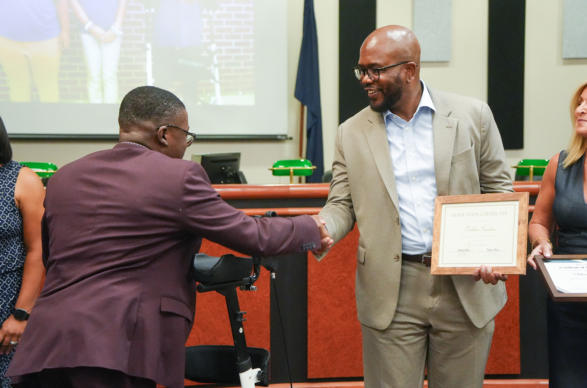 Two men shake hands in an indoor room, one holding a certificate. A woman stands nearby with a certificate. Behind them, a projector screen and flags are visible.