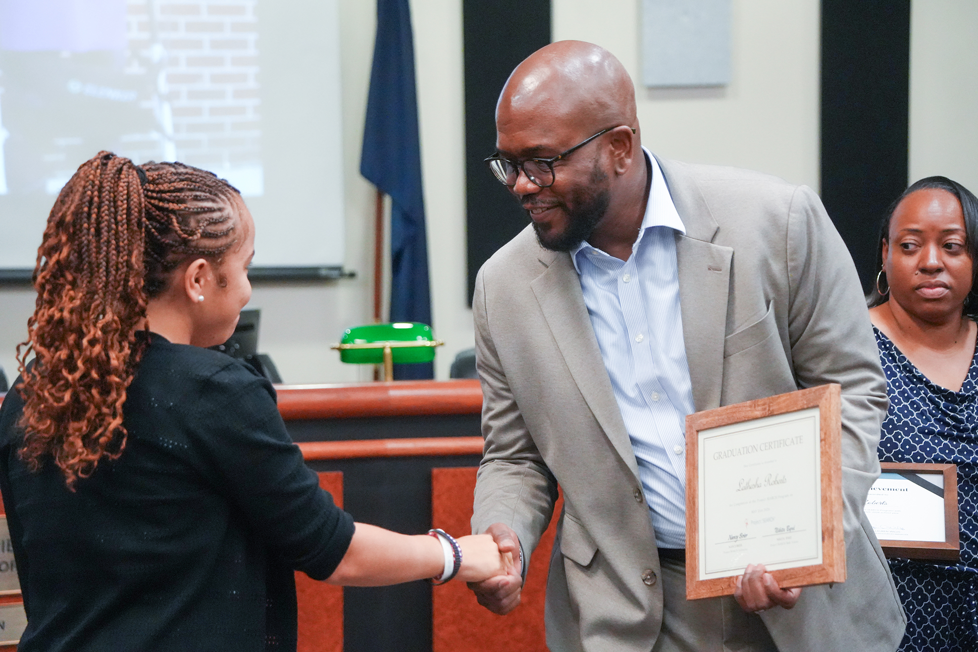 A person hands a framed certificate to a child with both hands. Behind them, a woman stands, and a flag hangs on the wall.