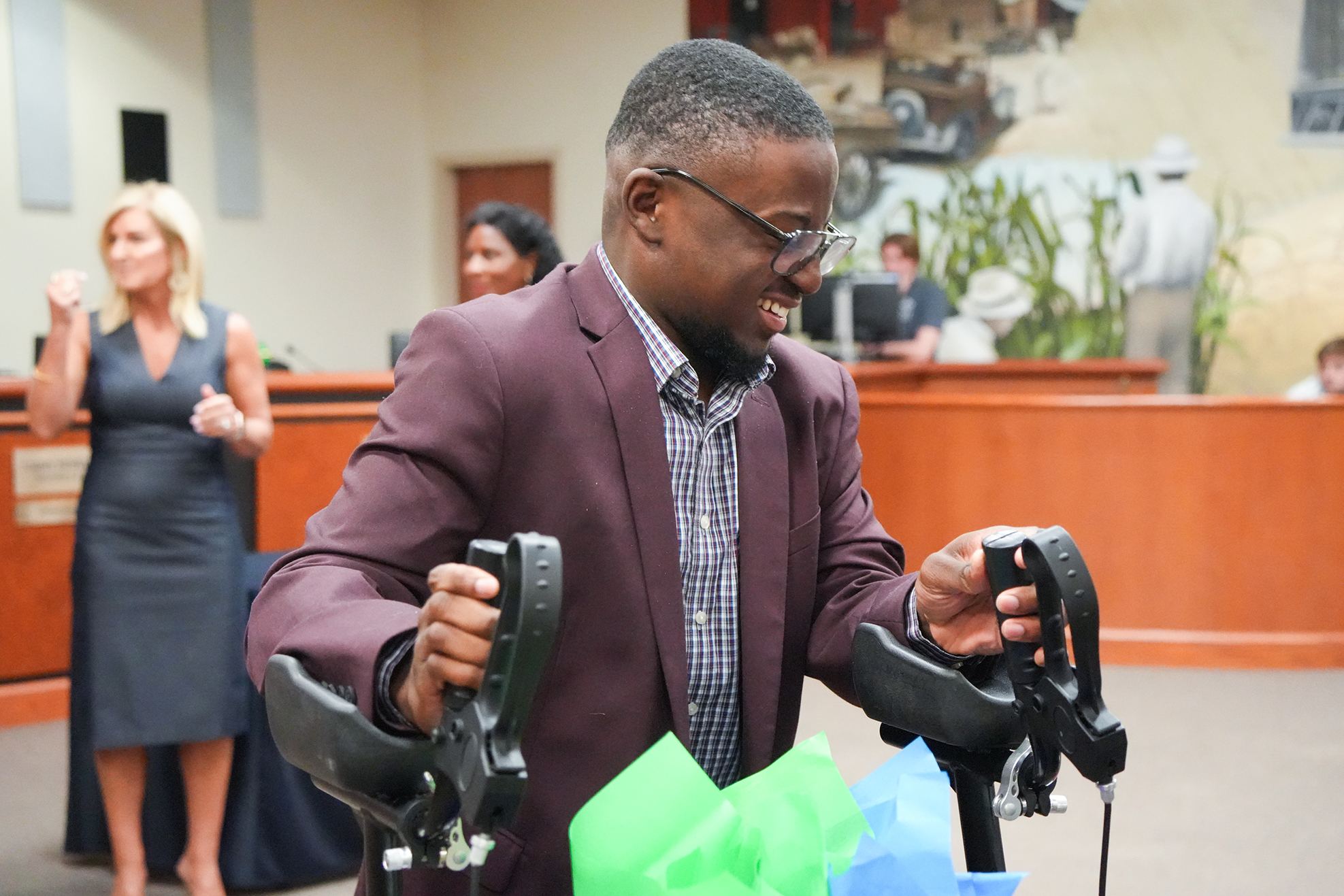 A man in a maroon suit and glasses holds a black object in both hands. A woman in a dress watches behind him. A wooden desk and greenery are in the background.