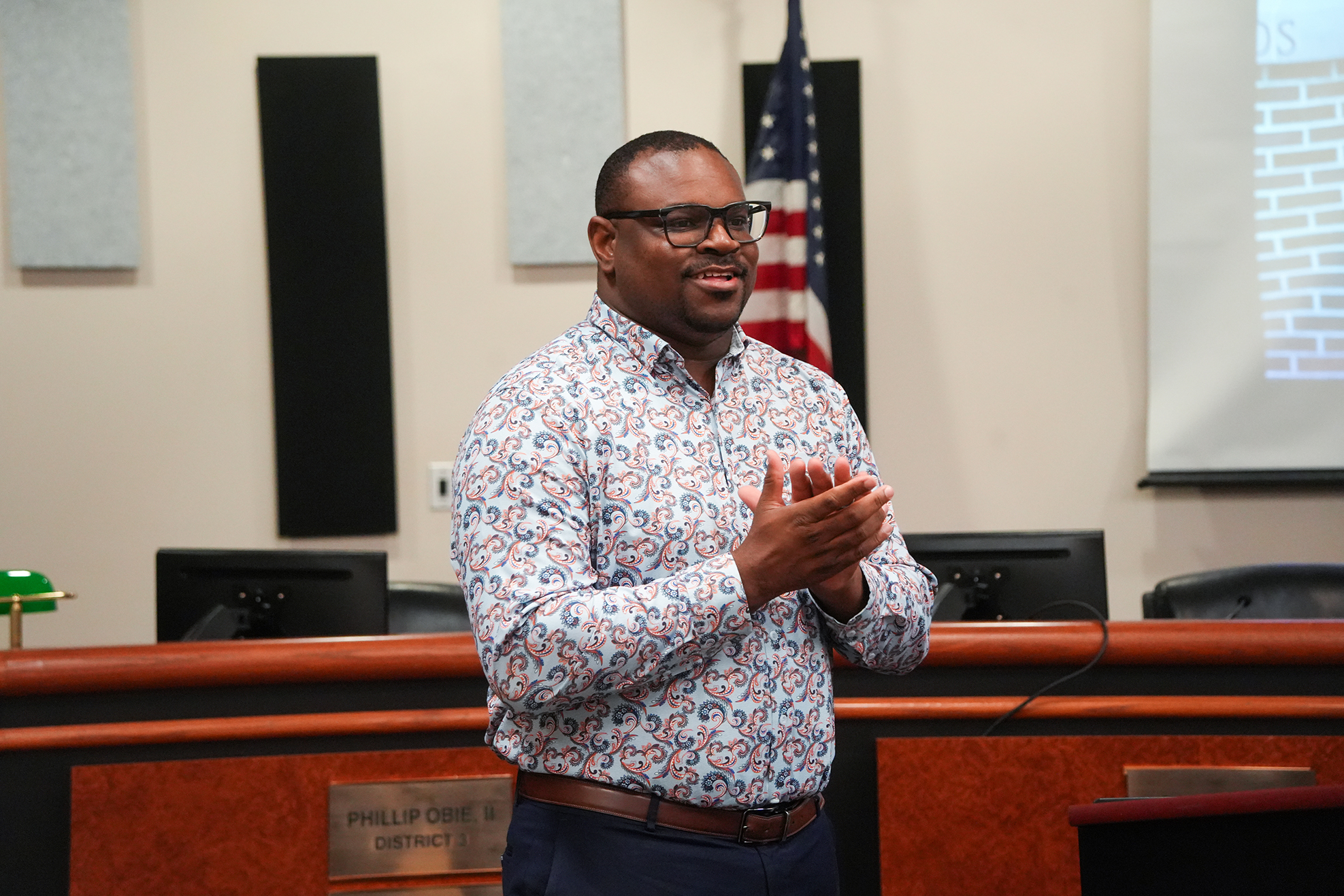 Person in a patterned shirt and glasses clapping in a room with a podium, flag, and screens.